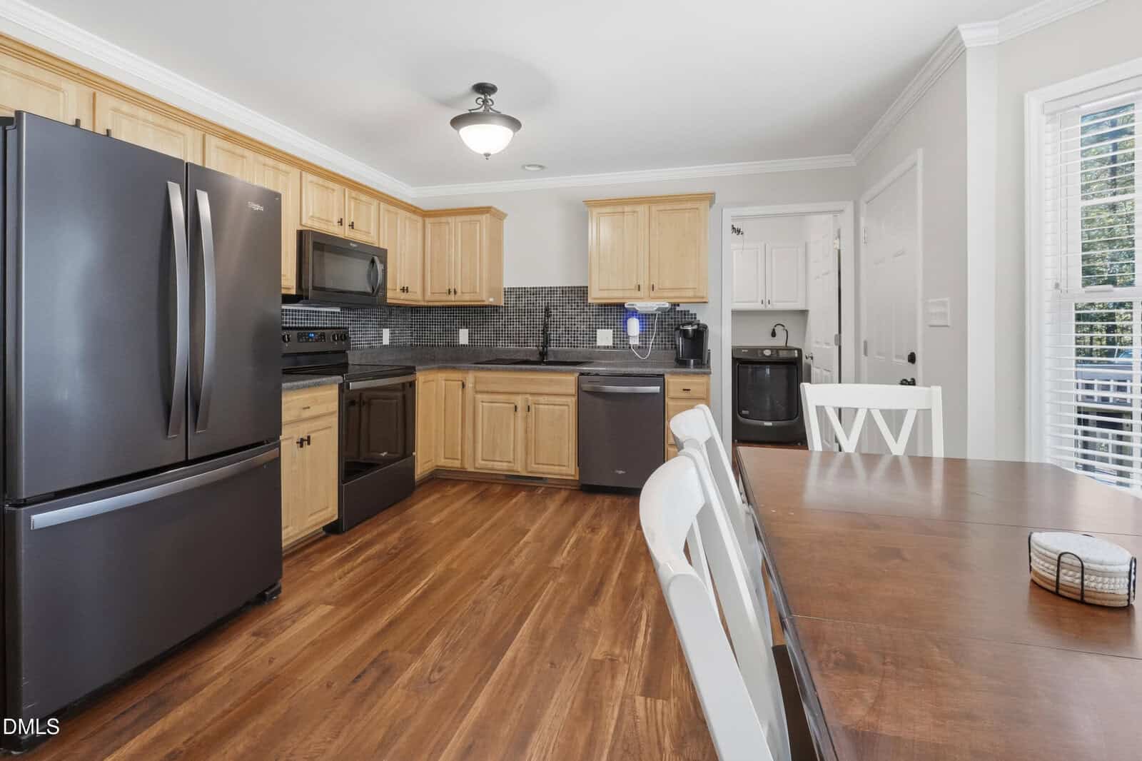 Modern kitchen with light wood cabinets, black appliances, and a dark tiled backsplash. A wooden dining table and chairs are in the foreground, with a laundry area visible in the back corner. Bright natural light enters from windows.