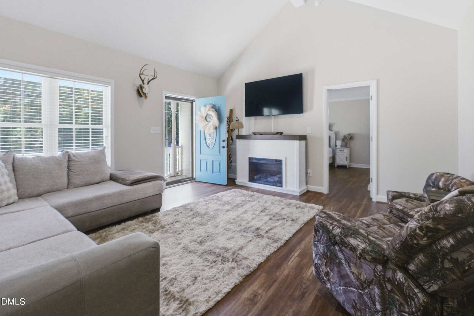 A living room with a gray sectional sofa, a large area rug, a mounted TV above a white fireplace, a camouflage armchair, and an open blue front door. Sunlight enters through large windows on the left.