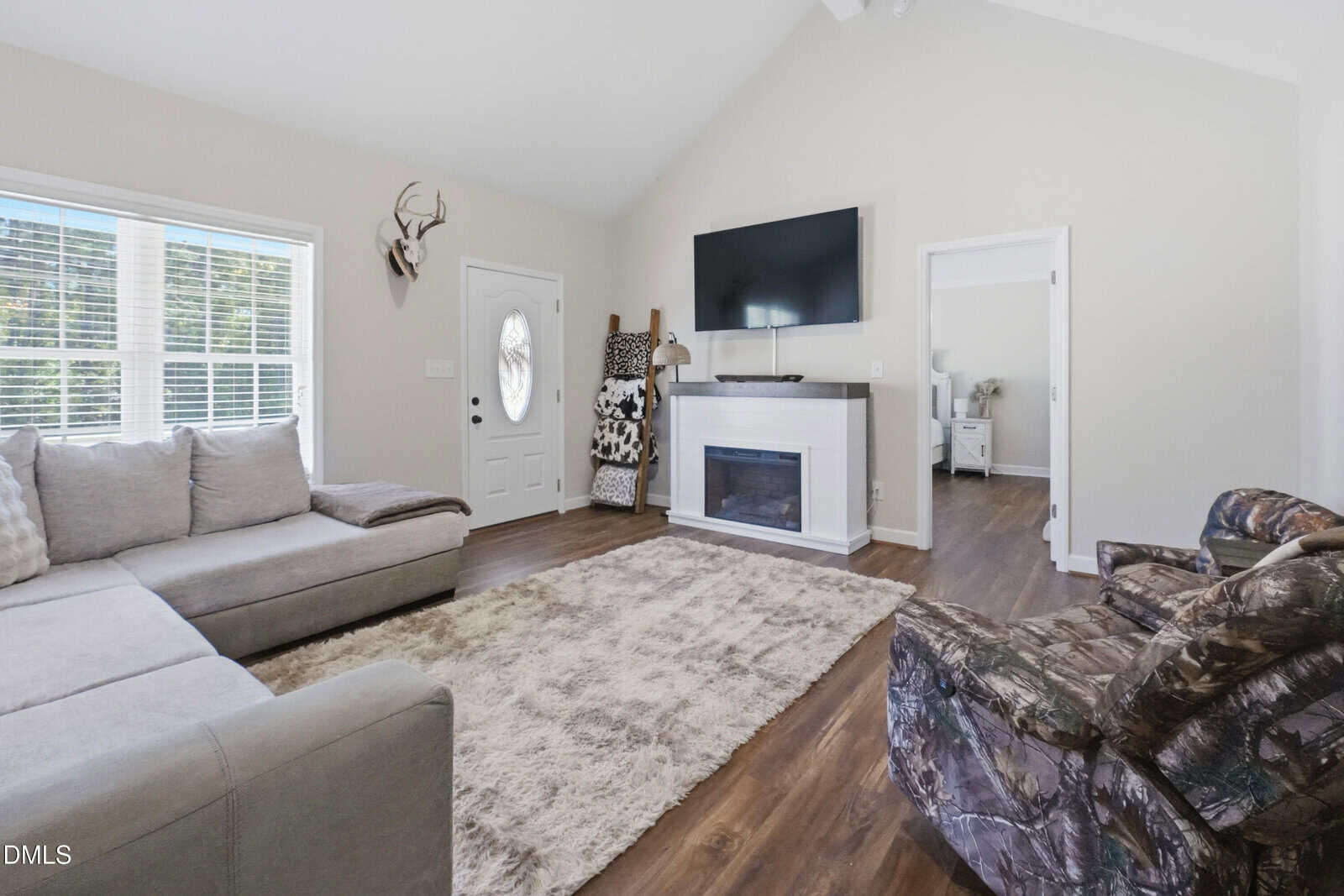 A living room with a sectional sofa, patterned recliner, wall-mounted TV above a fireplace, a beige rug on wood flooring, antlers on the wall, and a door with a circular window next to two large windows.