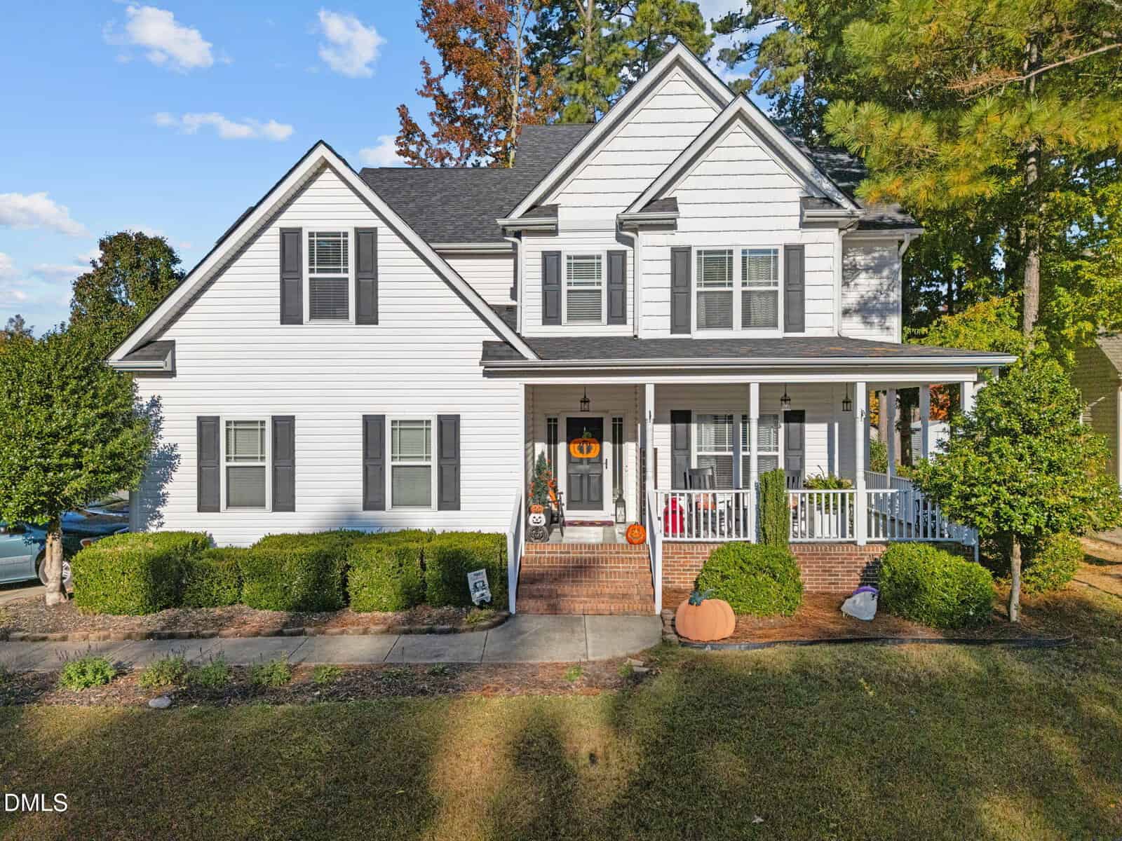 A two-story white house at 105 Torrance Court with black shutters and a covered front porch, decorated with fall items like pumpkins and a wreath. Bushes line the front, and trees stem beautifully in the background.