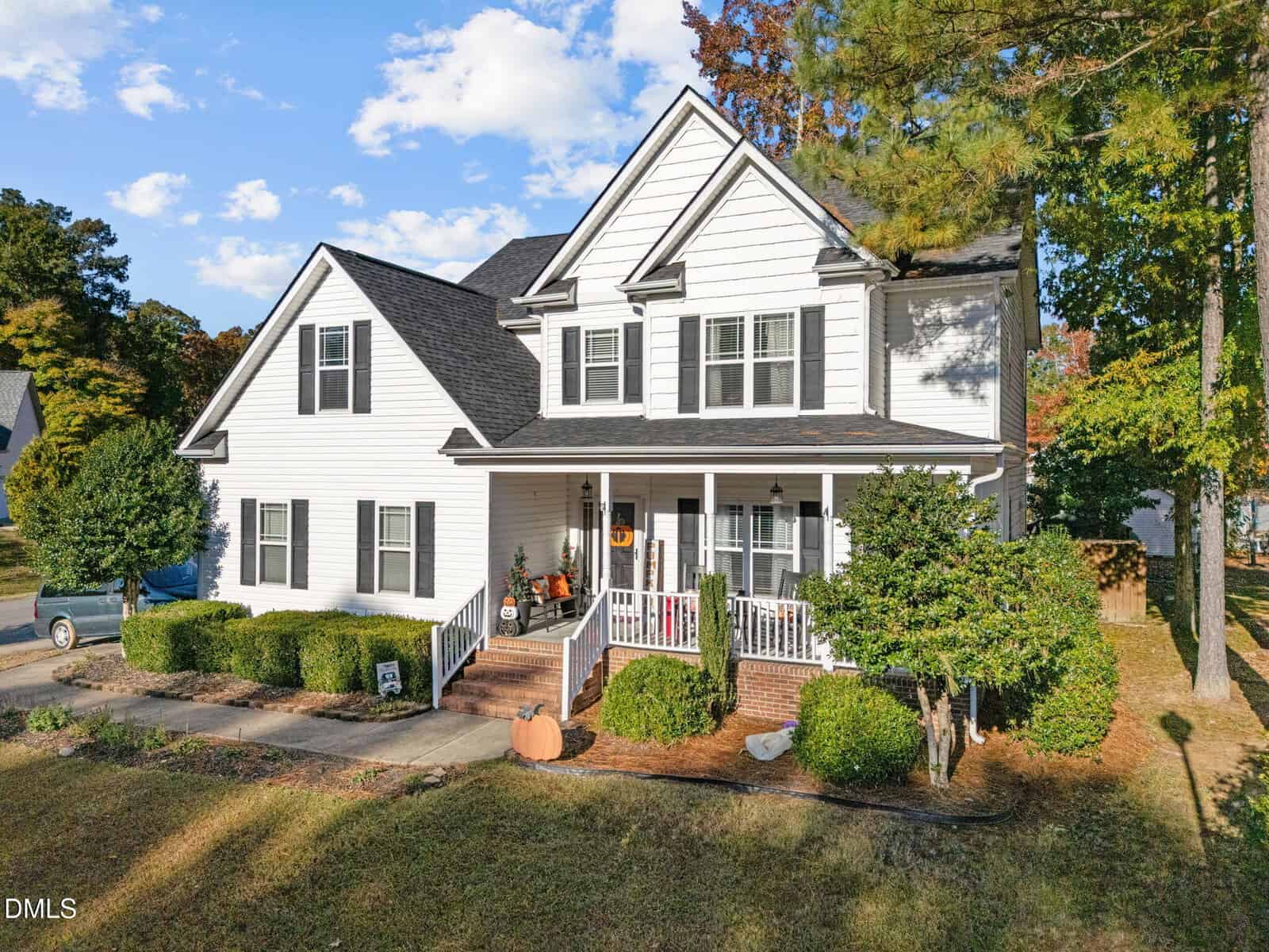 Two-story white house with black shutters and a covered front porch, featuring brick steps, potted plants, and a pumpkin. The yard has shrubs, mulch, a driveway, and is surrounded by trees on a sunny day.