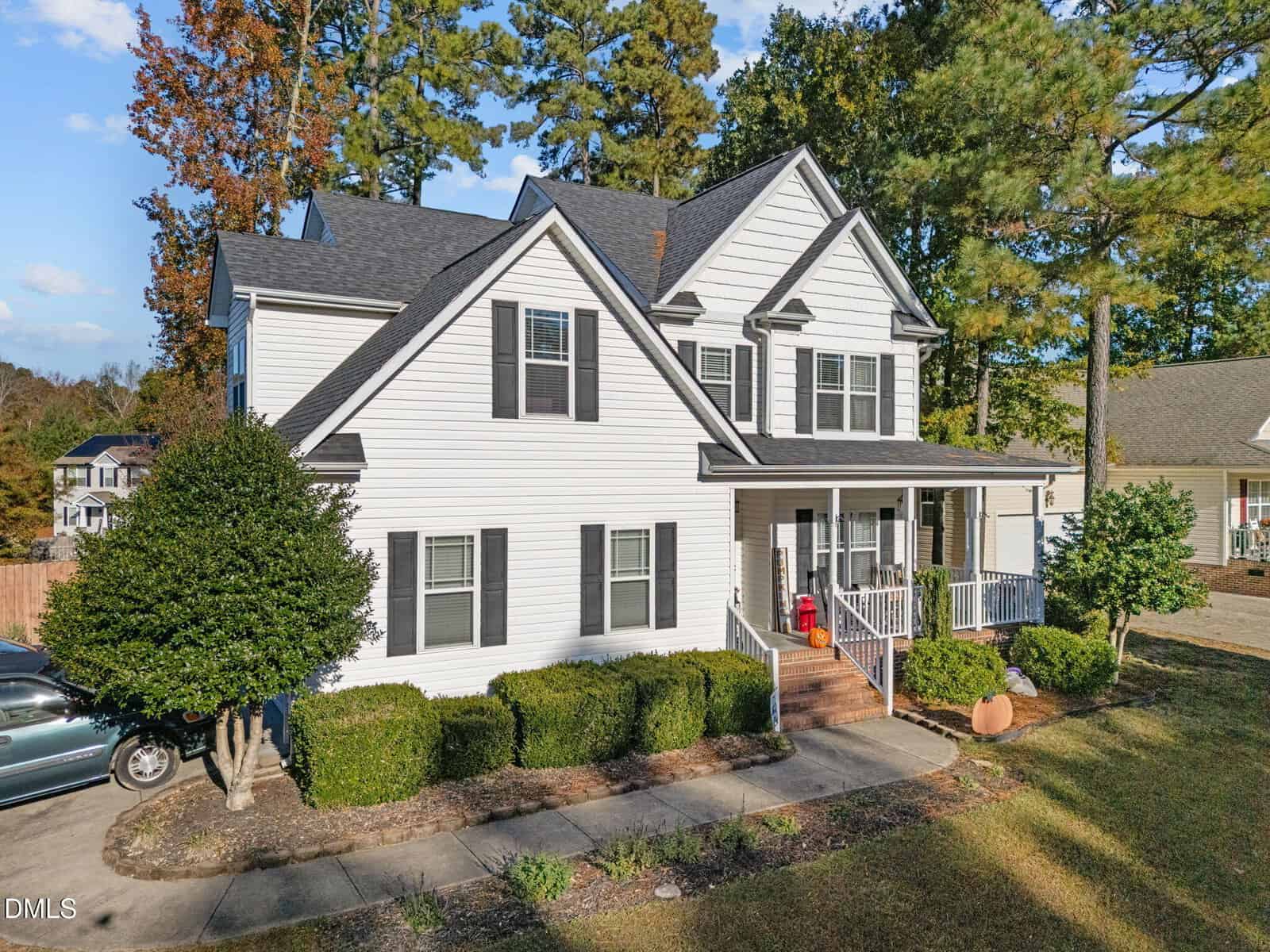 Two-story white house with black shutters, a front porch, and steps leading to the entrance. Shrubs line the walkway, and tall trees surround the property. A car is parked to the left, and the driveway is visible.