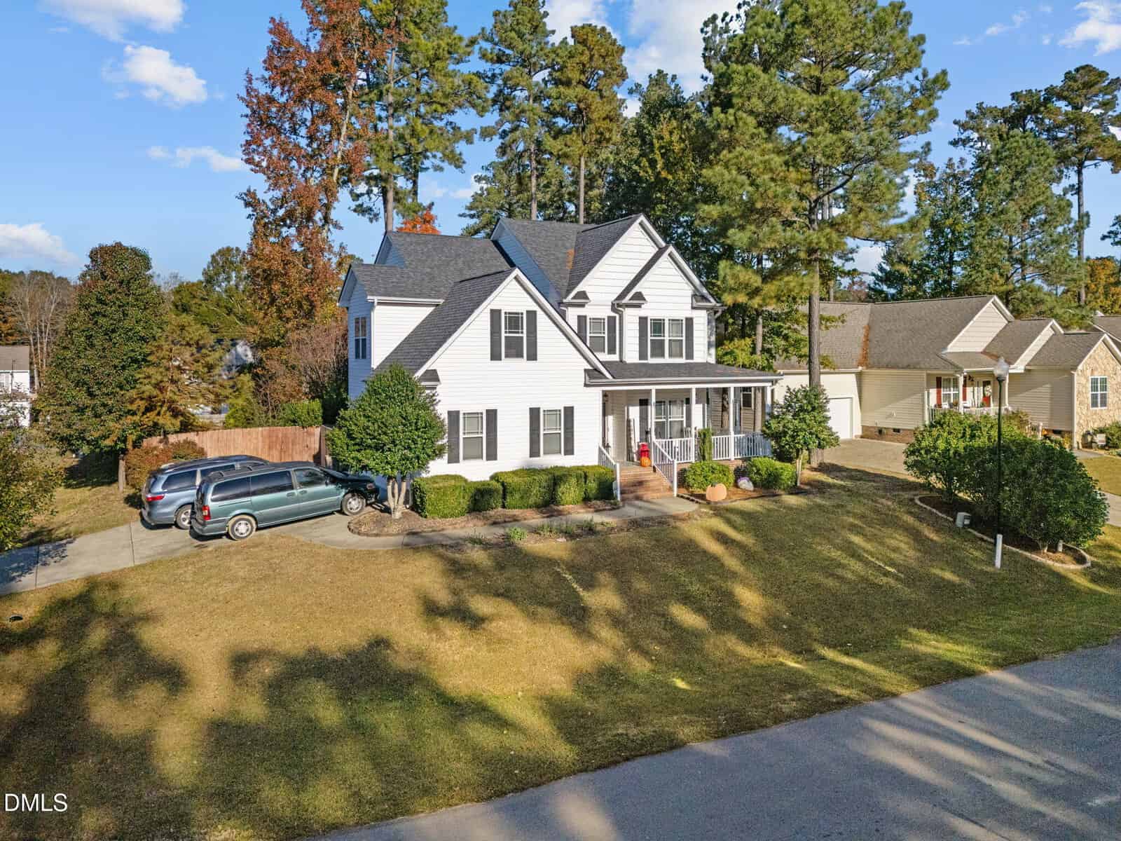 Two-story white house with a covered front porch, gray roof, and attached garage. Two cars are parked in the driveway. The yard is grassy with trimmed bushes and trees in the background. The house is in a suburban neighborhood.