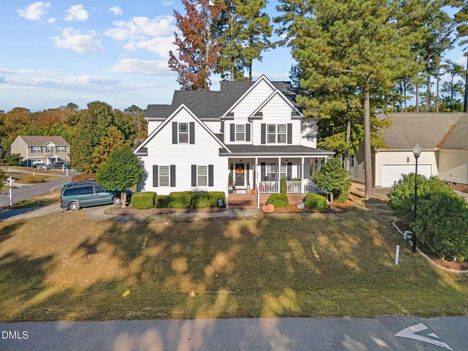 A two-story white house with black shutters and a covered front porch, situated on a corner lot with a few shrubs and trees. A dark minivan is parked in the driveway, and neighboring houses are visible in the background.