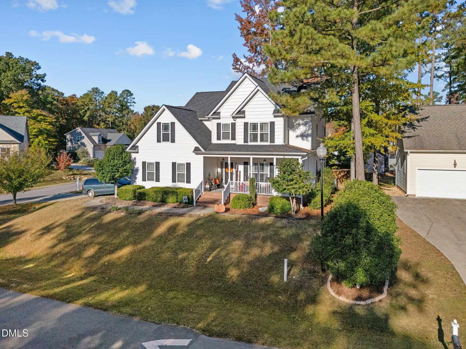 A two-story white suburban house with black shutters and a covered front porch, surrounded by neatly trimmed bushes and a lawn. A driveway and detached garage are on the right, with trees in the background.