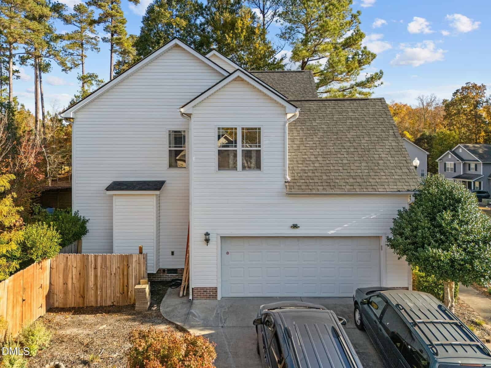 Two-story white house with gray shingle roof, double garage, and small front yard with a fence on one side. Two cars are parked in the driveway, and trees are visible in the background.