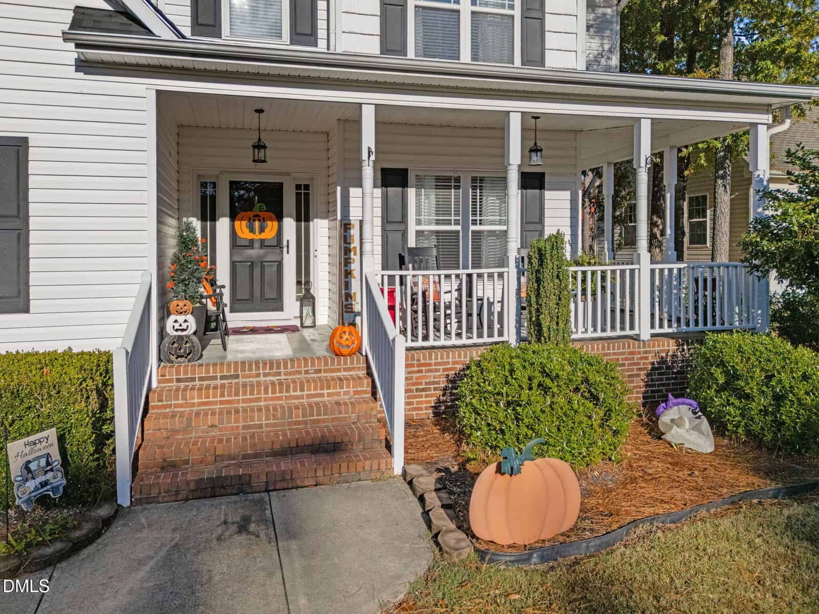 A white house with a brick porch decorated for Halloween, including pumpkins, jack-o'-lanterns, a small Halloween tree, and a ghost figure on the front lawn. Bushes line the walkway to the steps.