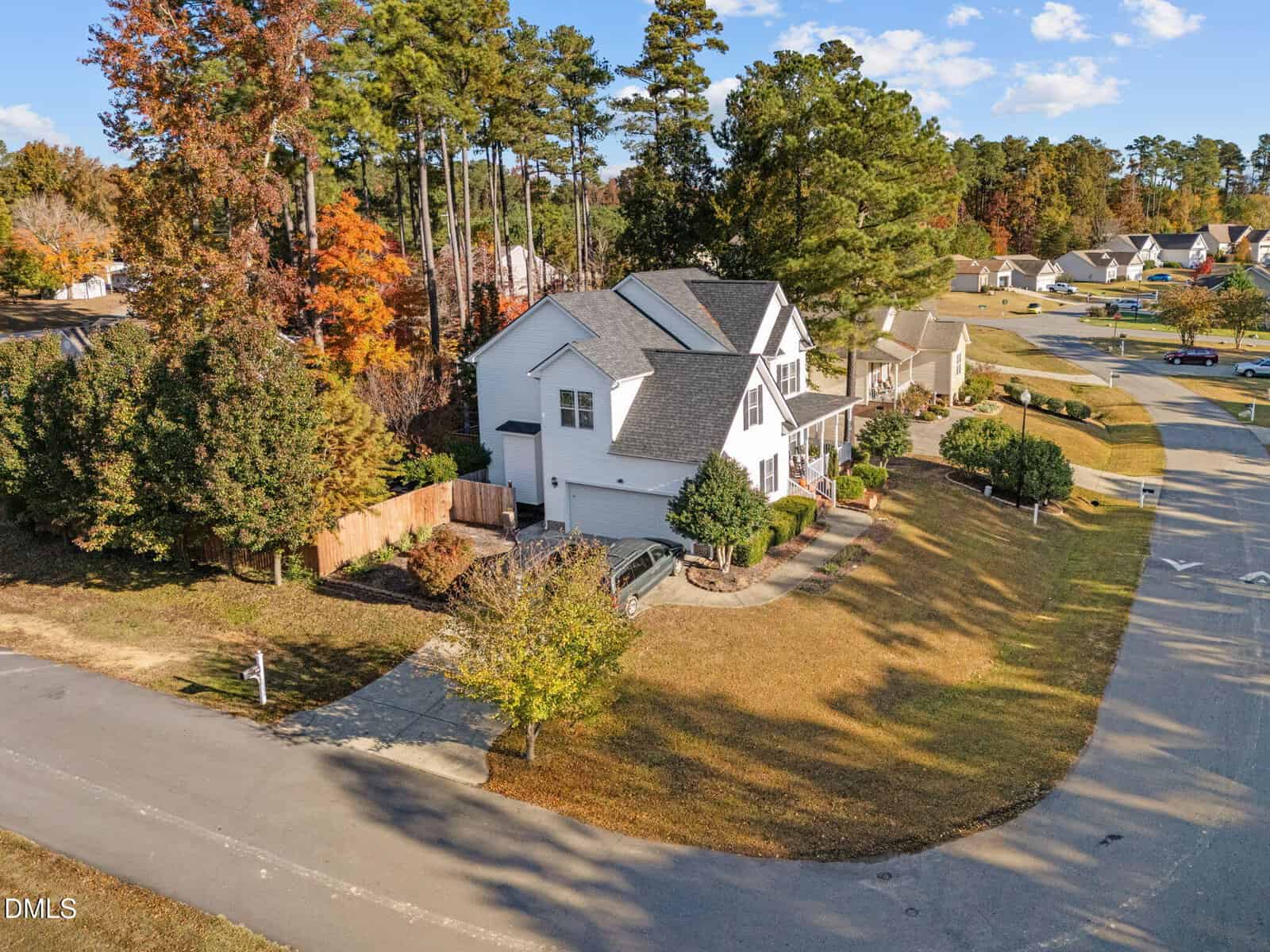 A two-story white house with a gray roof sits on a corner lot with a fenced backyard, driveway, and well-maintained lawn. Trees with autumn foliage surround the property and neighboring homes line the street.