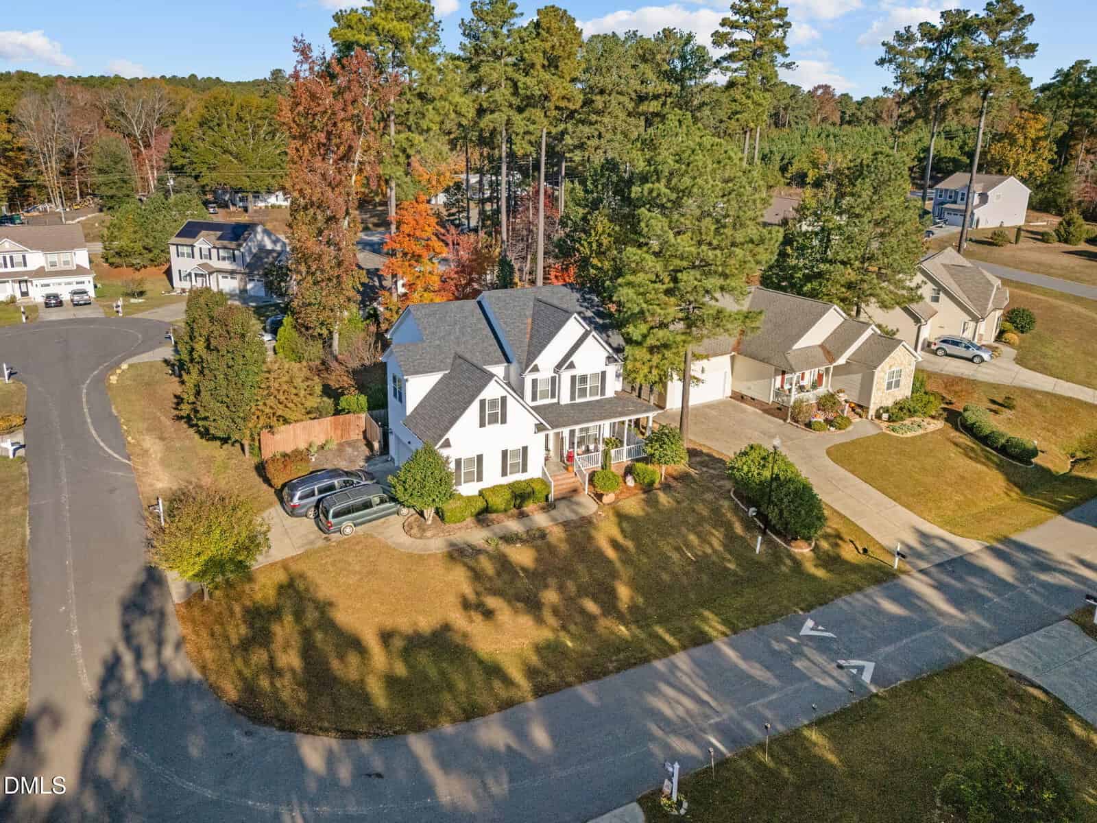 Aerial view of a suburban neighborhood with houses, driveways, cars, trees, and lawns on a sunny day. One white house is centered, surrounded by greenery and a curving street.