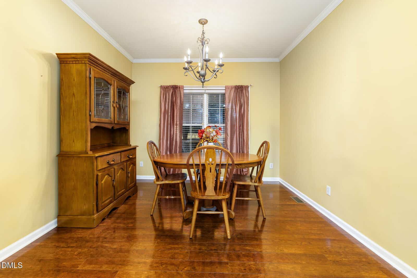 A dining room with a wooden table, four chairs, a wooden hutch on the left, a chandelier overhead, and a window with pink curtains at the back. The floor is hardwood and the walls are painted light yellow.