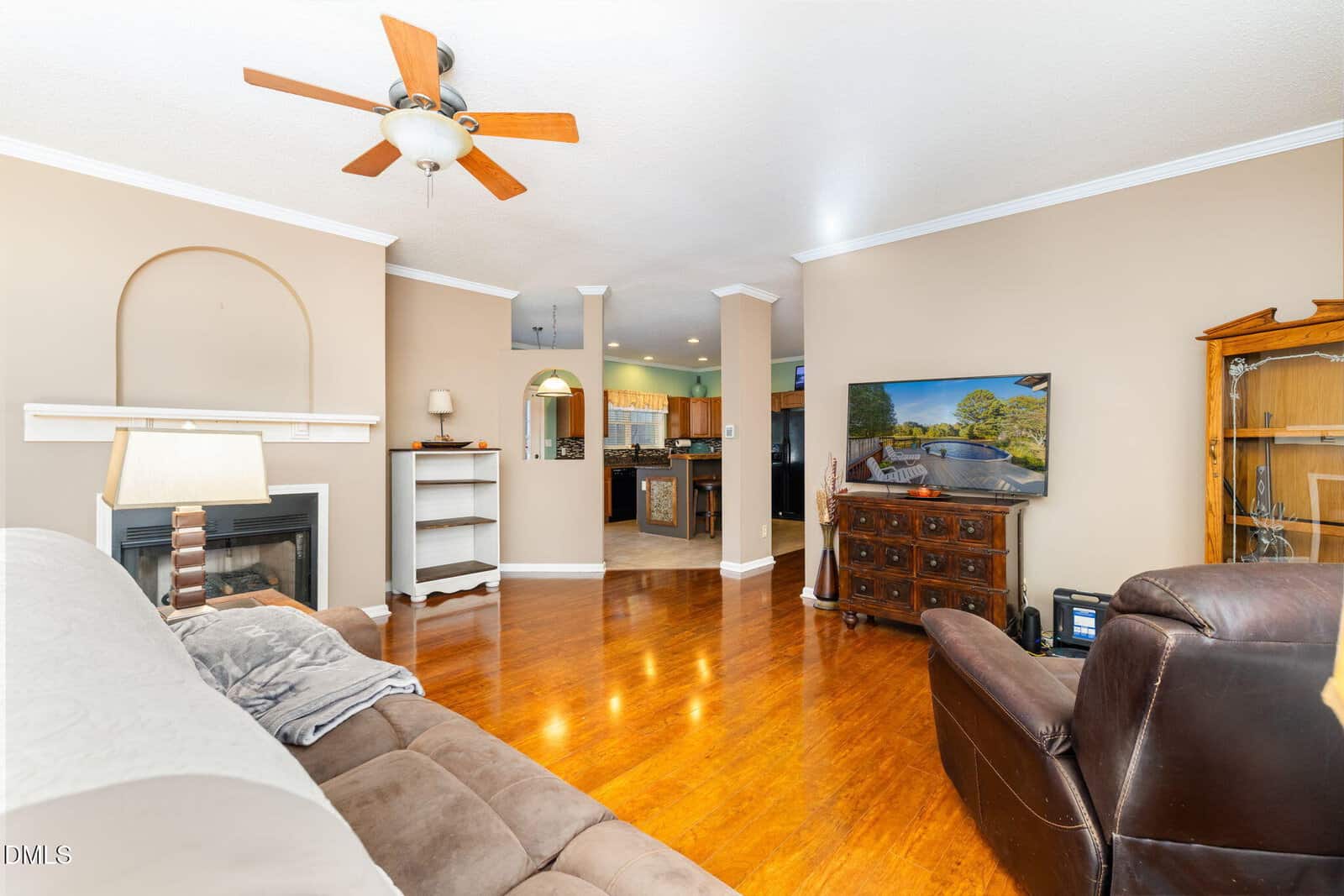 A living room with wood flooring, a ceiling fan, a brown sofa, and a leather recliner. A TV sits on a wooden dresser, facing the seating area. The room opens into a kitchen with visible countertops and cabinets.