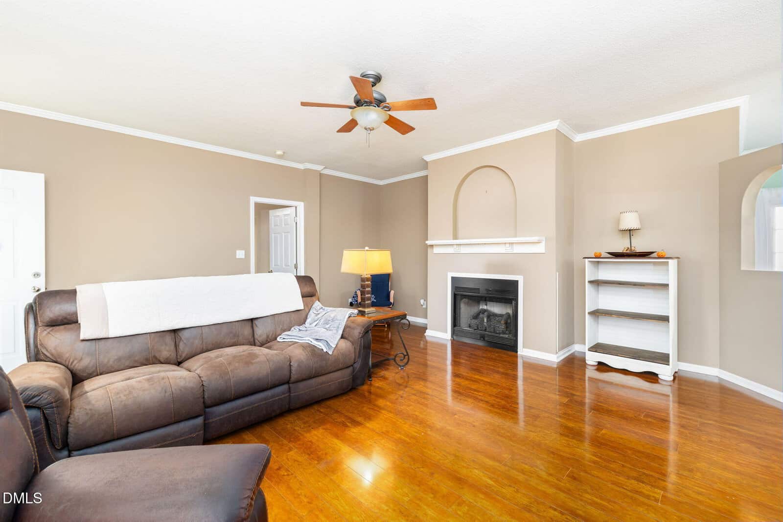 A living room with tan walls, a brown leather sectional sofa, wooden floors, a ceiling fan, a fireplace with a white mantel, a small bookshelf, a table lamp, and a doorway visible in the background.