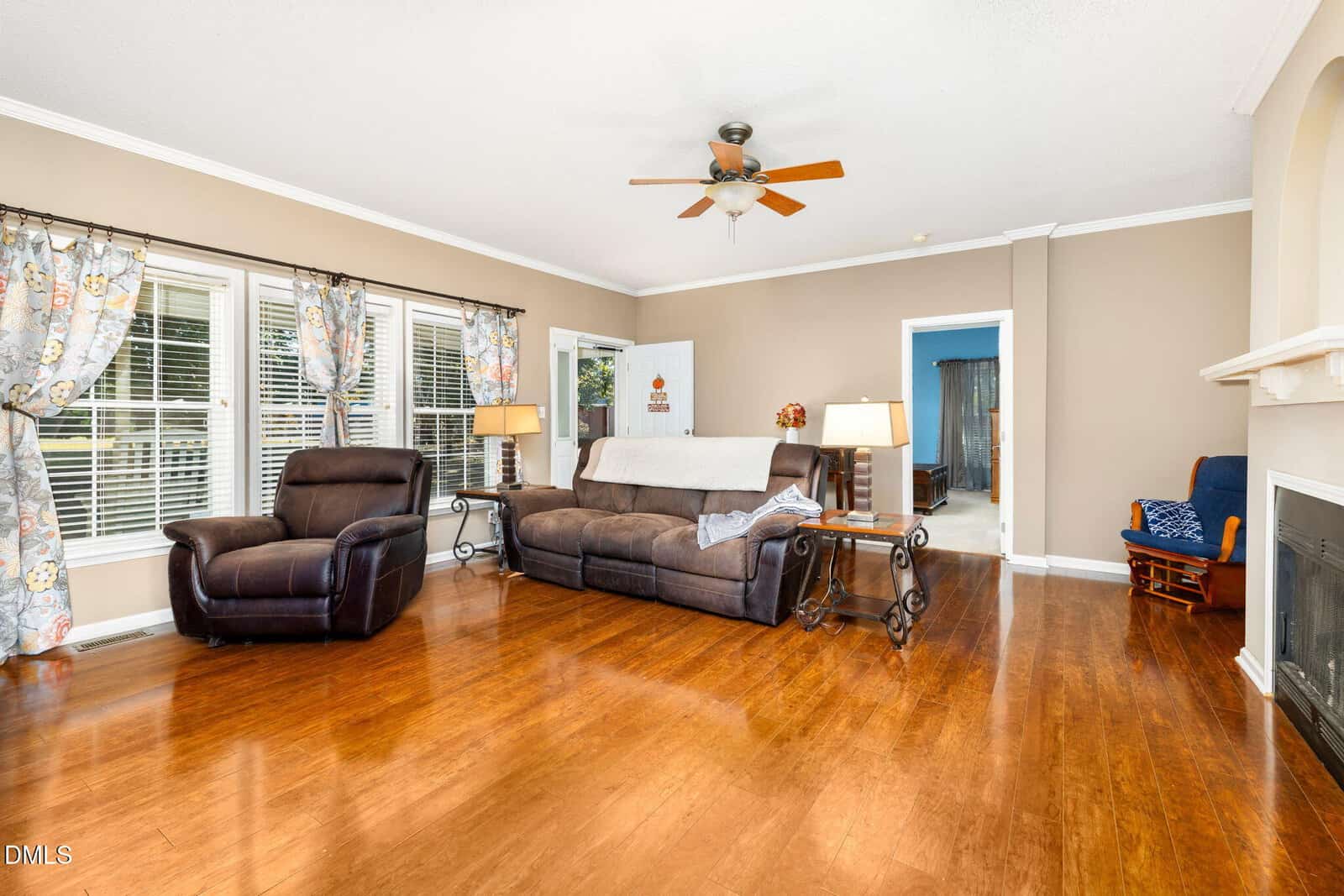 A spacious living room with brown hardwood floors, a brown leather armchair and sofa, floral curtains on large windows, a ceiling fan, and a fireplace with a white mantle. A bedroom is visible through an open doorway.