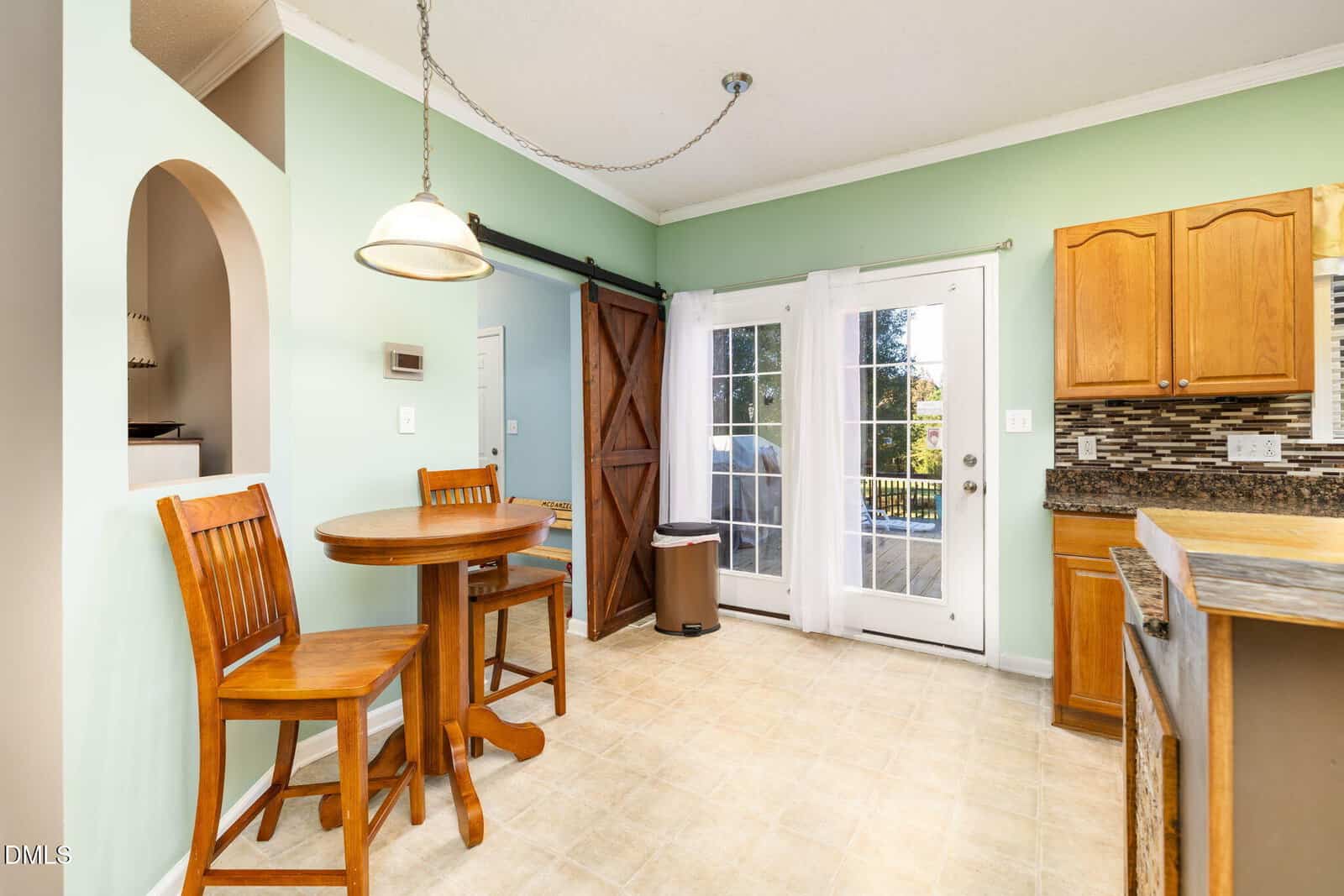 A small dining area with a round wooden table and two chairs, pale green walls, a sliding barn door, double glass doors leading outside, and part of a kitchen counter with wooden cabinets.