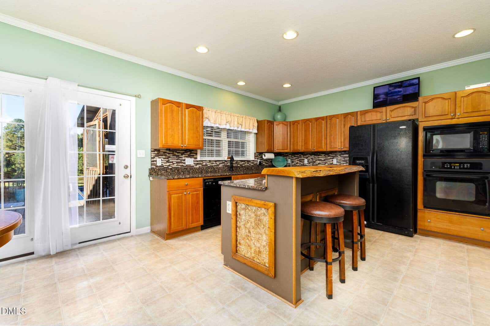 A kitchen with light brown cabinets, black appliances, a granite countertop, a raised breakfast bar with two stools, tile flooring, and green walls. French doors lead outside, and a window is above the sink.