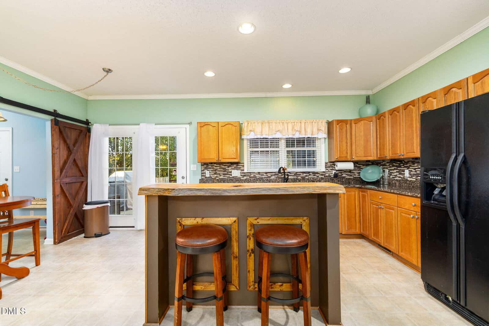A kitchen with wooden cabinets, a two-tier island with two brown barstools, black appliances including a refrigerator, a double sink under a window, and French doors leading outside. Walls are light green with crown molding.