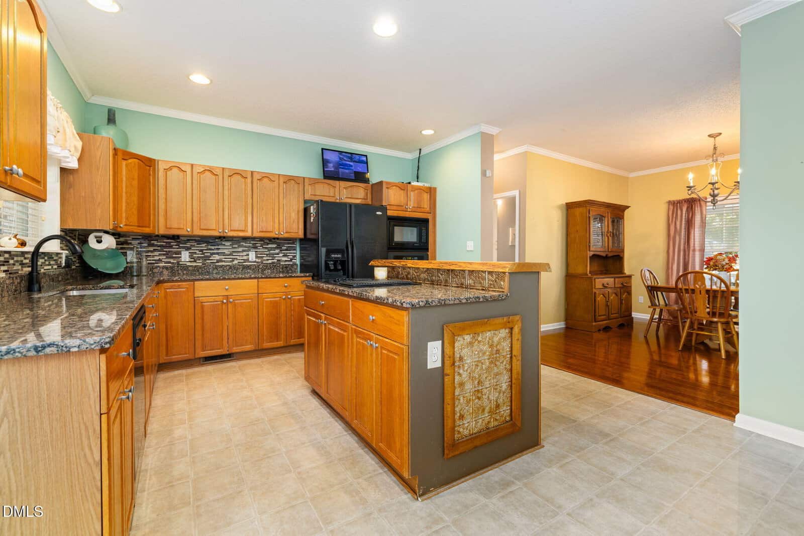 Kitchen with wooden cabinets, granite countertops, black appliances, and tile flooring opens into a dining area with a wooden hutch, chandelier, and wood floor. Walls are painted light green and yellow.