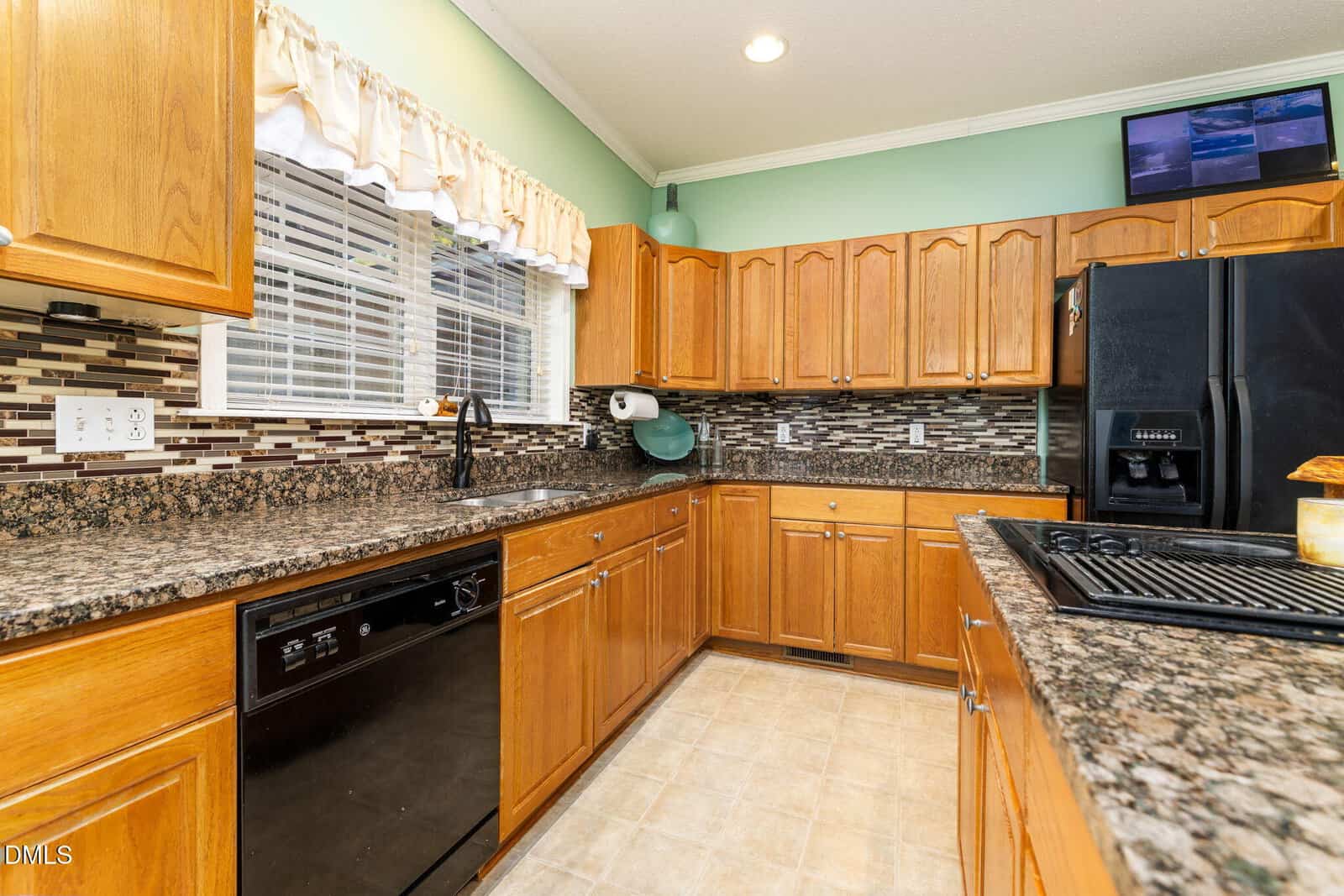 A kitchen with wooden cabinets, black appliances, granite countertops, a tiled backsplash, a double sink under a window with a white valance, and a TV mounted above the cabinets.