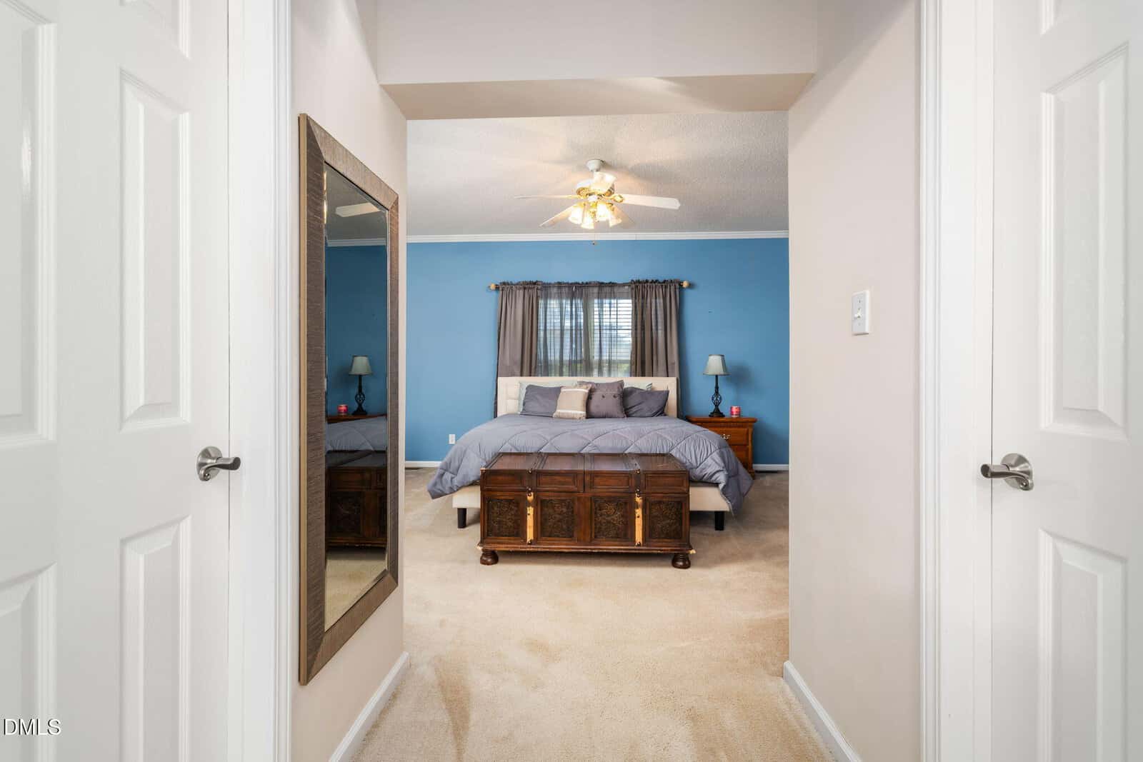 A hallway with white doors leads to a bedroom with blue walls, a ceiling fan, a bed with gray bedding, two nightstands with lamps, and a large mirror on the left wall.