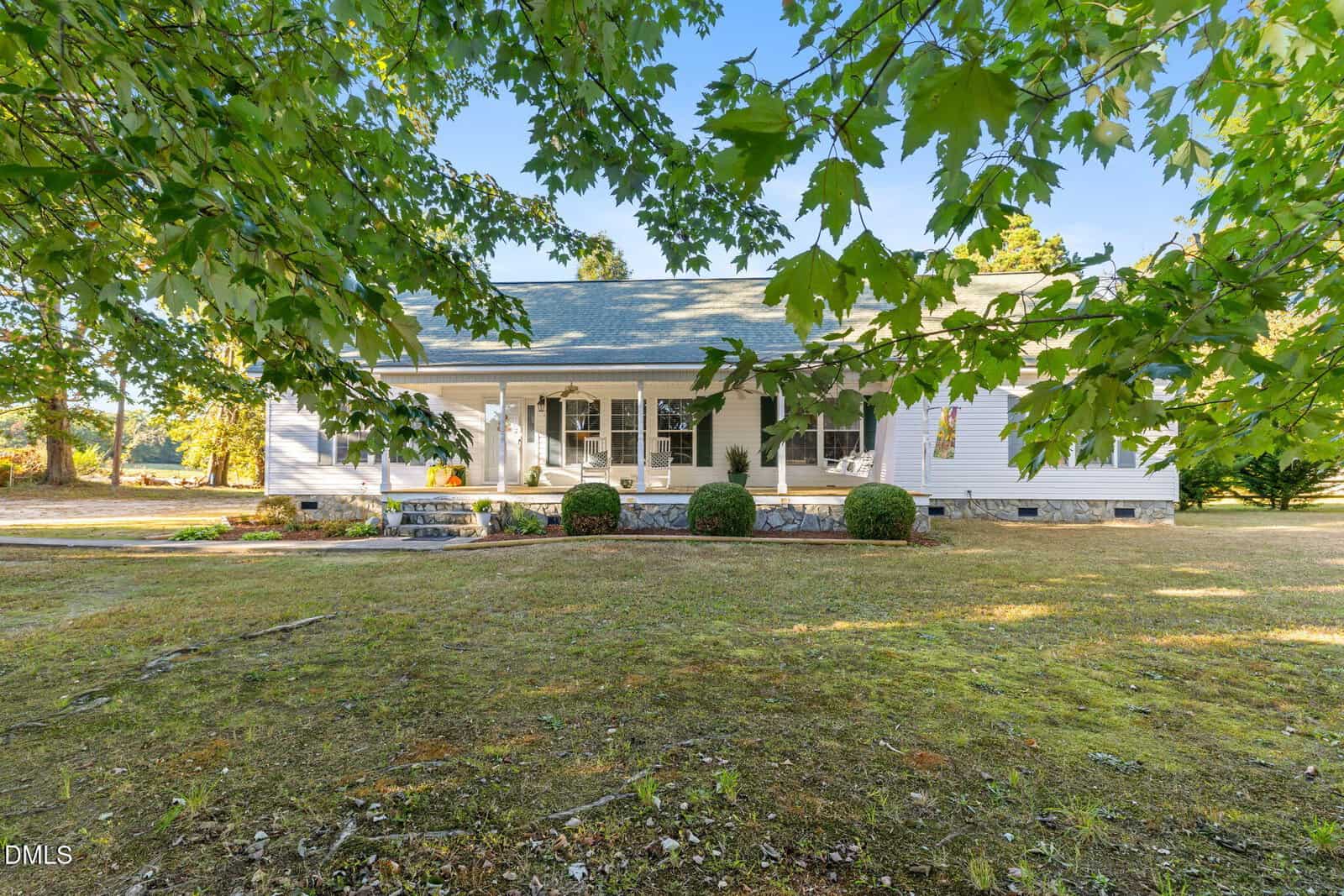 A white single-story house with a covered front porch, several chairs, and bushes along the stone foundation, viewed through green leafy branches in the foreground. The yard has patchy grass and scattered trees.