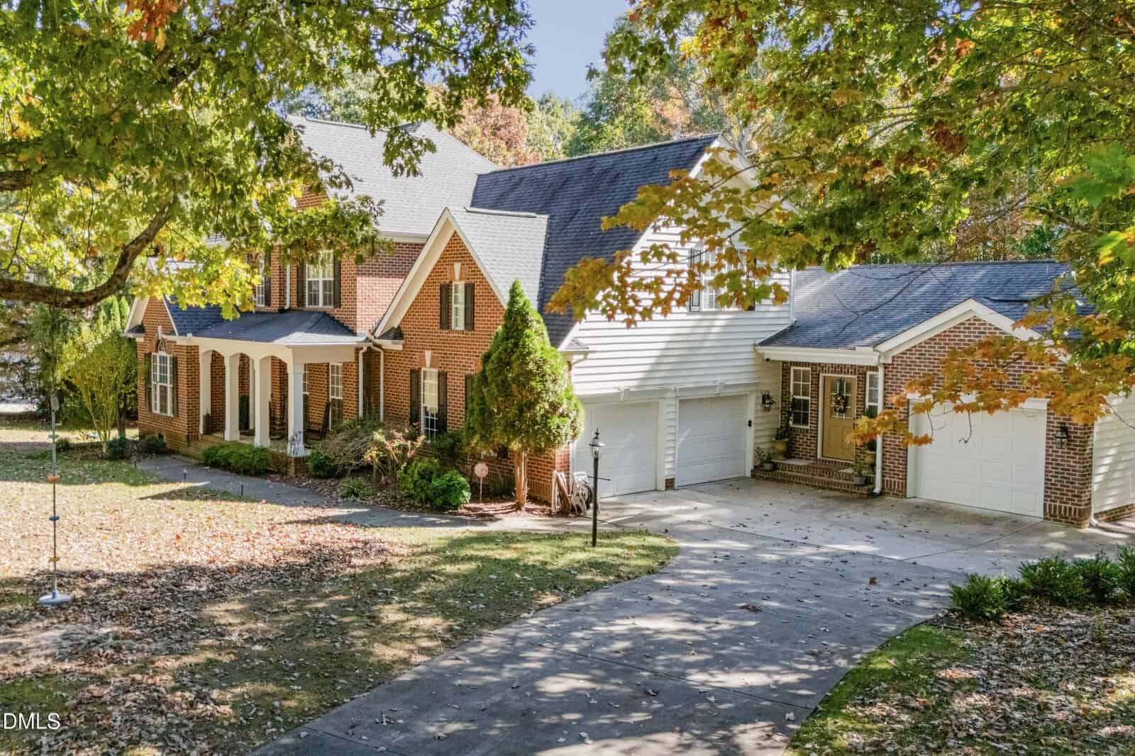 Two-story red brick house with white trim, dormer windows, and an attached two-car garage located at 185 Kensington Drive in Youngsville. A curved concrete driveway leads to the garage, and mature trees with autumn leaves surround the property.