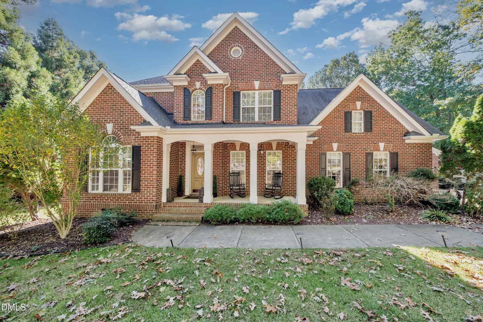 Two-story brick house with white trim, black shutters, and a covered front porch featuring two rocking chairs. The house is surrounded by trees, bushes, and a lawn with scattered autumn leaves. Blue sky with clouds overhead.