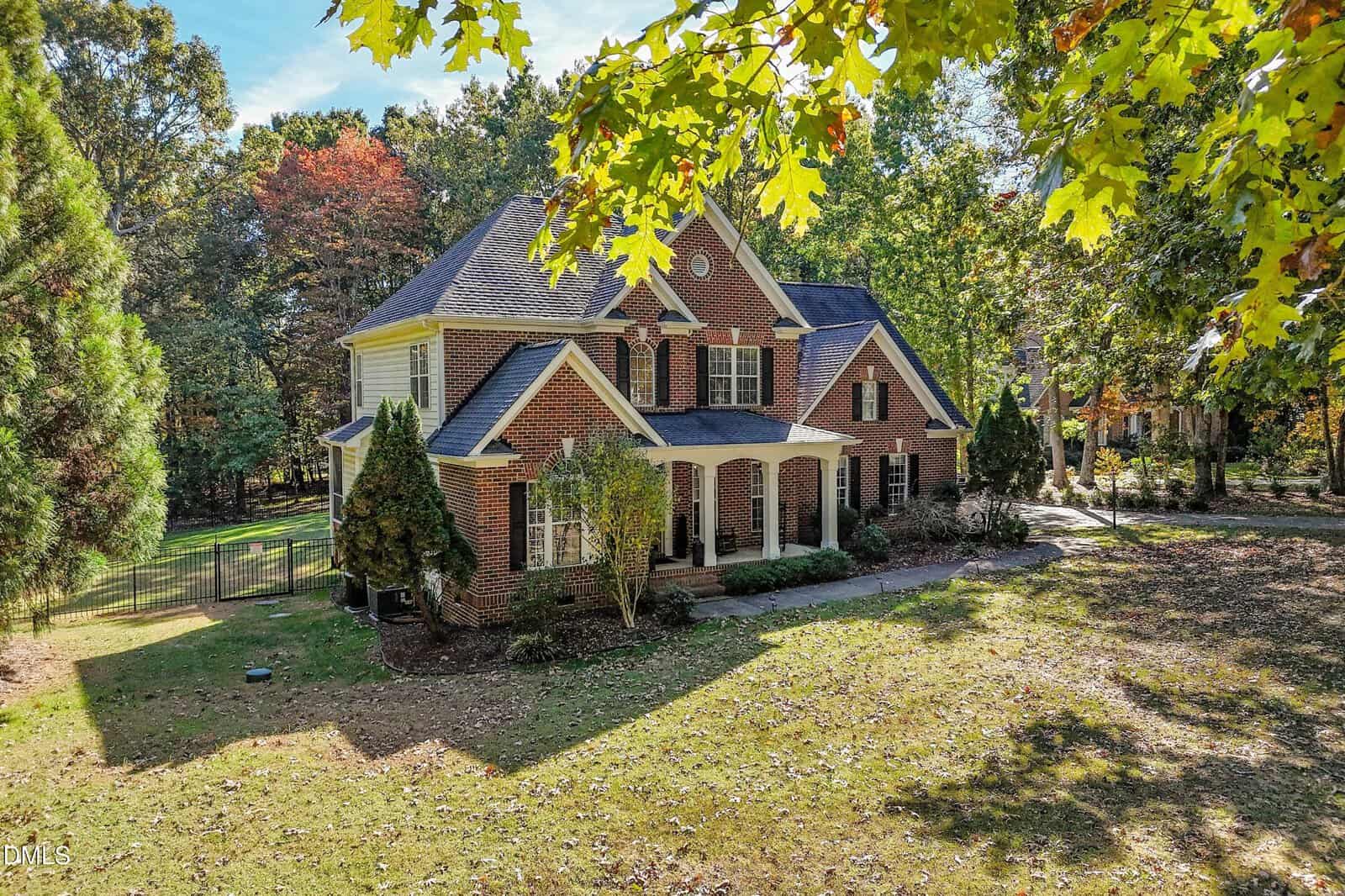 Two-story brick house with white columns, dark shutters, and a covered front porch, surrounded by trees and a grassy yard. The scene is bright with sunlight and scattered autumn leaves.