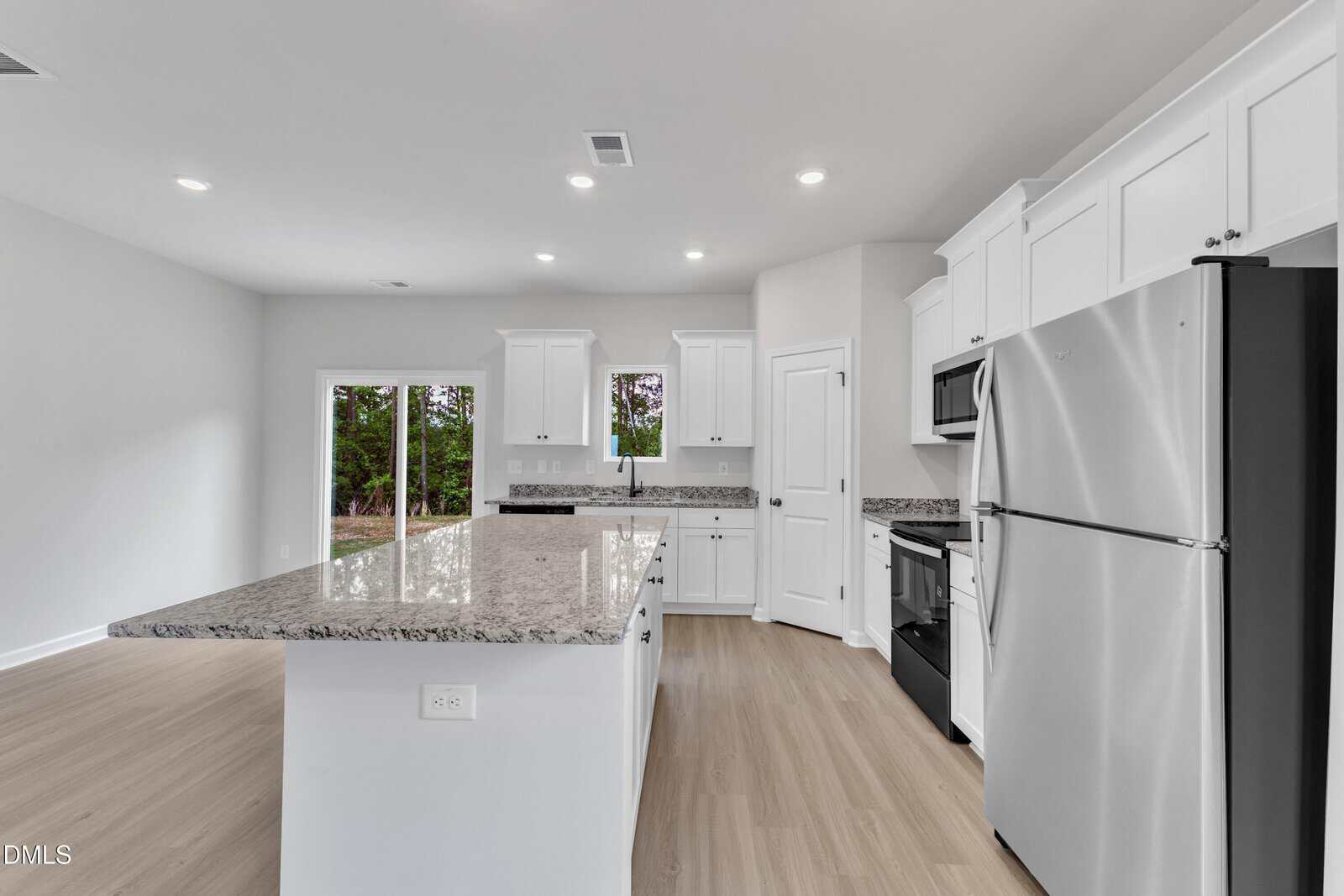 Modern kitchen with white cabinets, stainless steel appliances, granite countertops, a large island, light wood flooring, and a pantry door. Sliding glass doors in the background open to a view of trees.