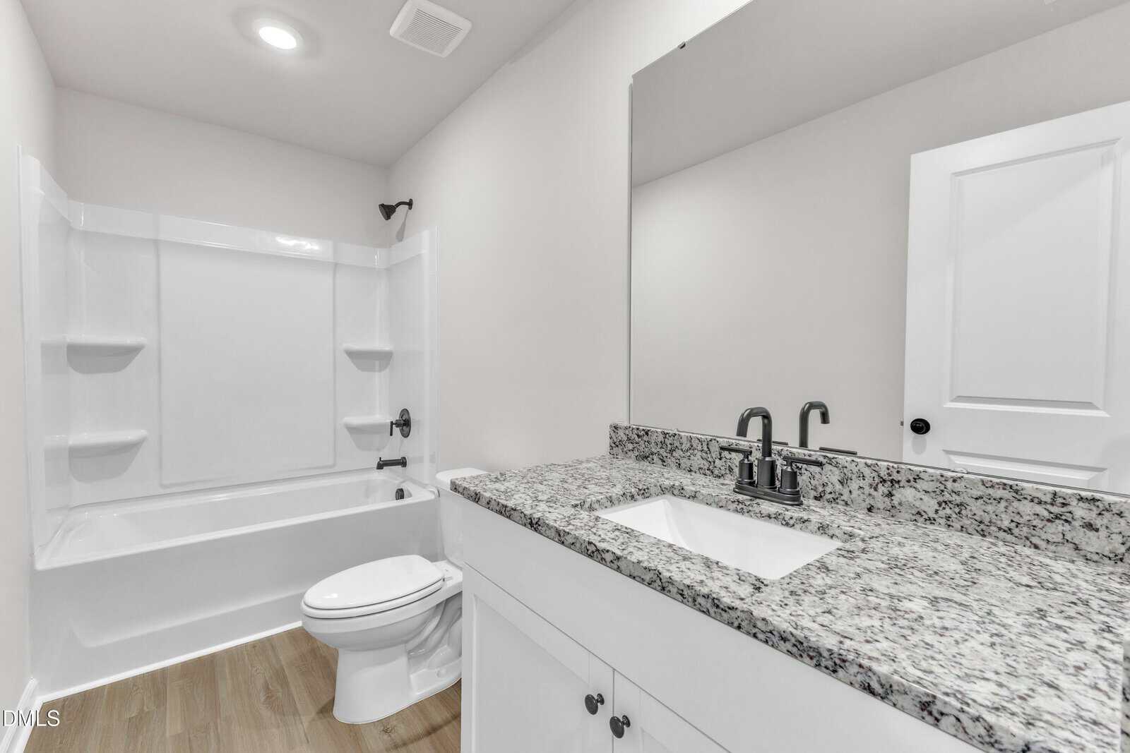 A modern bathroom with a white bathtub and shower, a toilet, a white vanity with a granite countertop, an undermount sink, a large mirror, and light wood-look flooring.