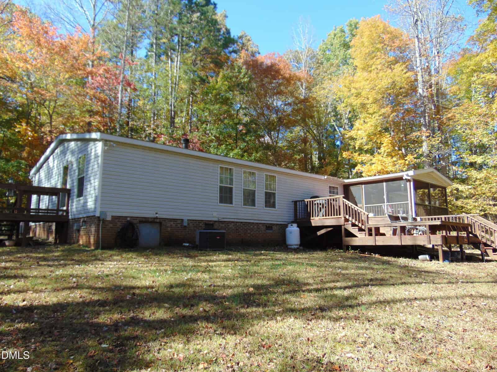 A single-story manufactured home with white siding sits on a grassy lawn. The house has multiple windows and a wooden deck with a screened porch, surrounded by tall trees with autumn foliage.