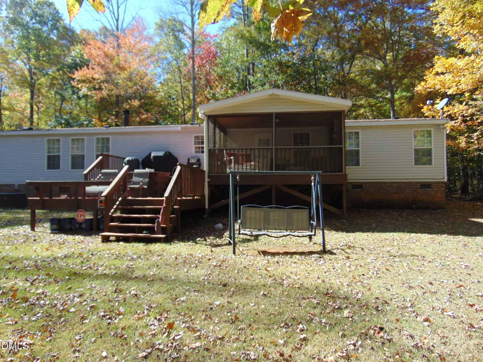 A single-story house with light siding and a brick foundation is surrounded by trees with autumn foliage. The backyard features two wooden decks, a covered porch, a grill, and a metal swing on a grassy lawn.