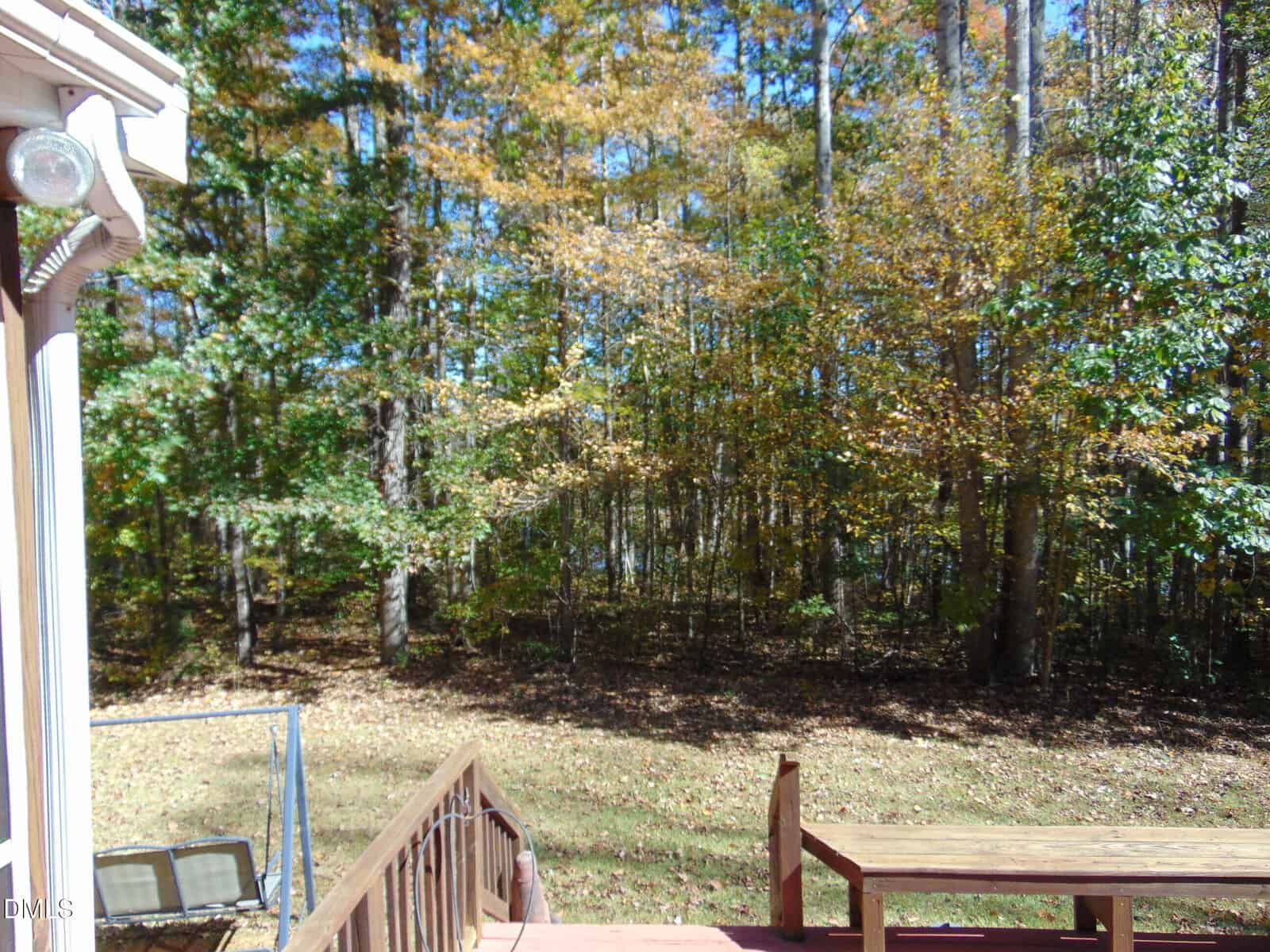 A wooden deck with railing and a bench overlooks a grassy backyard bordered by dense trees with green and yellow leaves. Part of a house and a metal-framed swing are visible on the left.