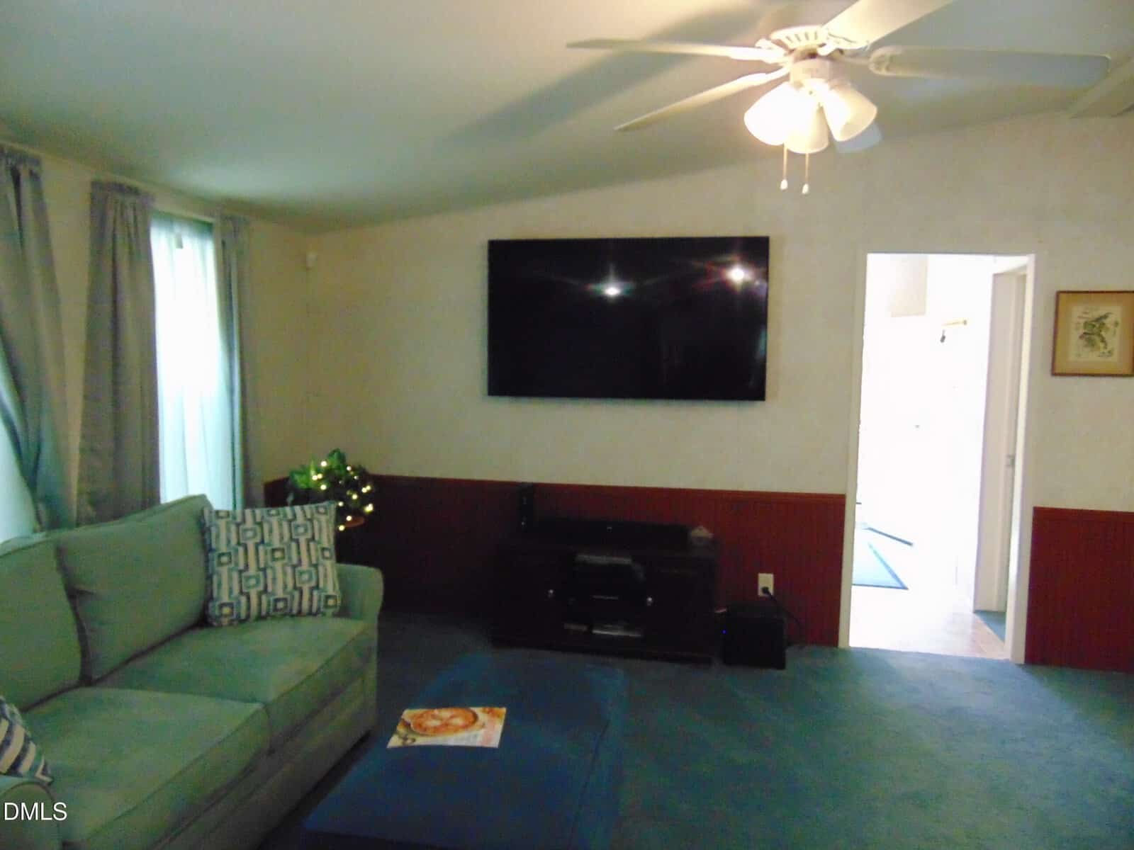 A living room with a green sectional sofa, a blue ottoman, a TV mounted on the wall above a media console, light carpet, curtained windows, and a ceiling fan with lights. A door leads to another room.