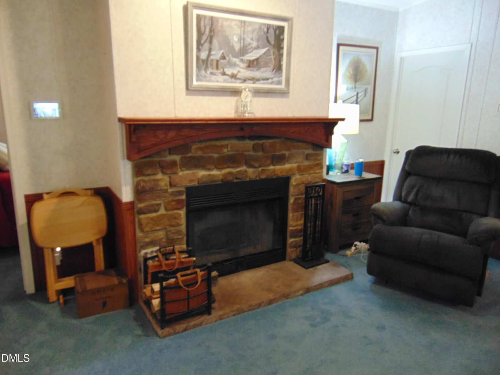 A stone fireplace with a wooden mantel in a living room, flanked by a black recliner chair and a small table with a lamp. Fireplace tools and firewood holder are placed on the hearth.