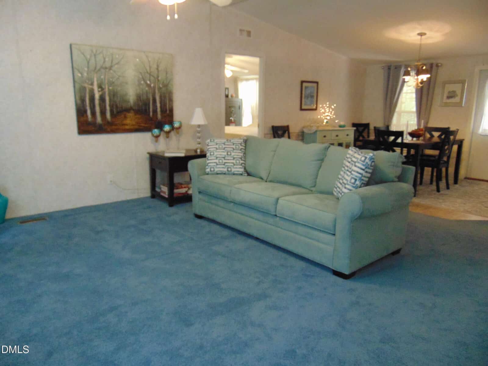 A living room with blue carpet, a light blue sofa, a side table with a lamp, and a wall painting of trees. In the background, a dining table with chairs is visible under a chandelier.