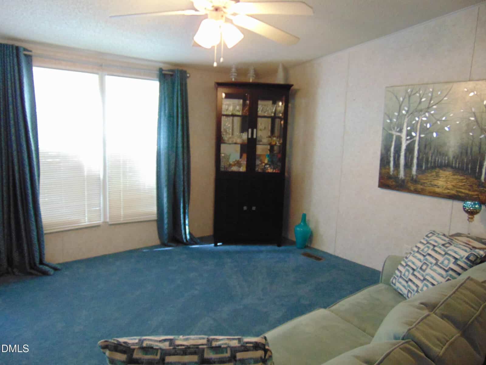 A living room with blue carpet, a ceiling fan, a window with blue curtains, a dark wood display cabinet, a large painting of trees on the wall, and a green couch with patterned pillows.