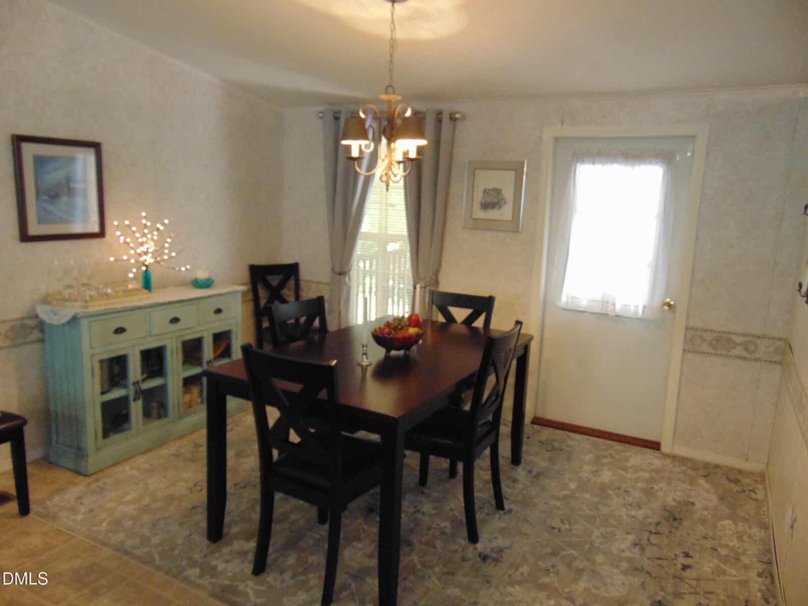 A dining room with a dark wooden table and six black chairs, a light-colored patterned rug, a blue sideboard with decor items, chandelier lighting, and a door with a sheer curtain.