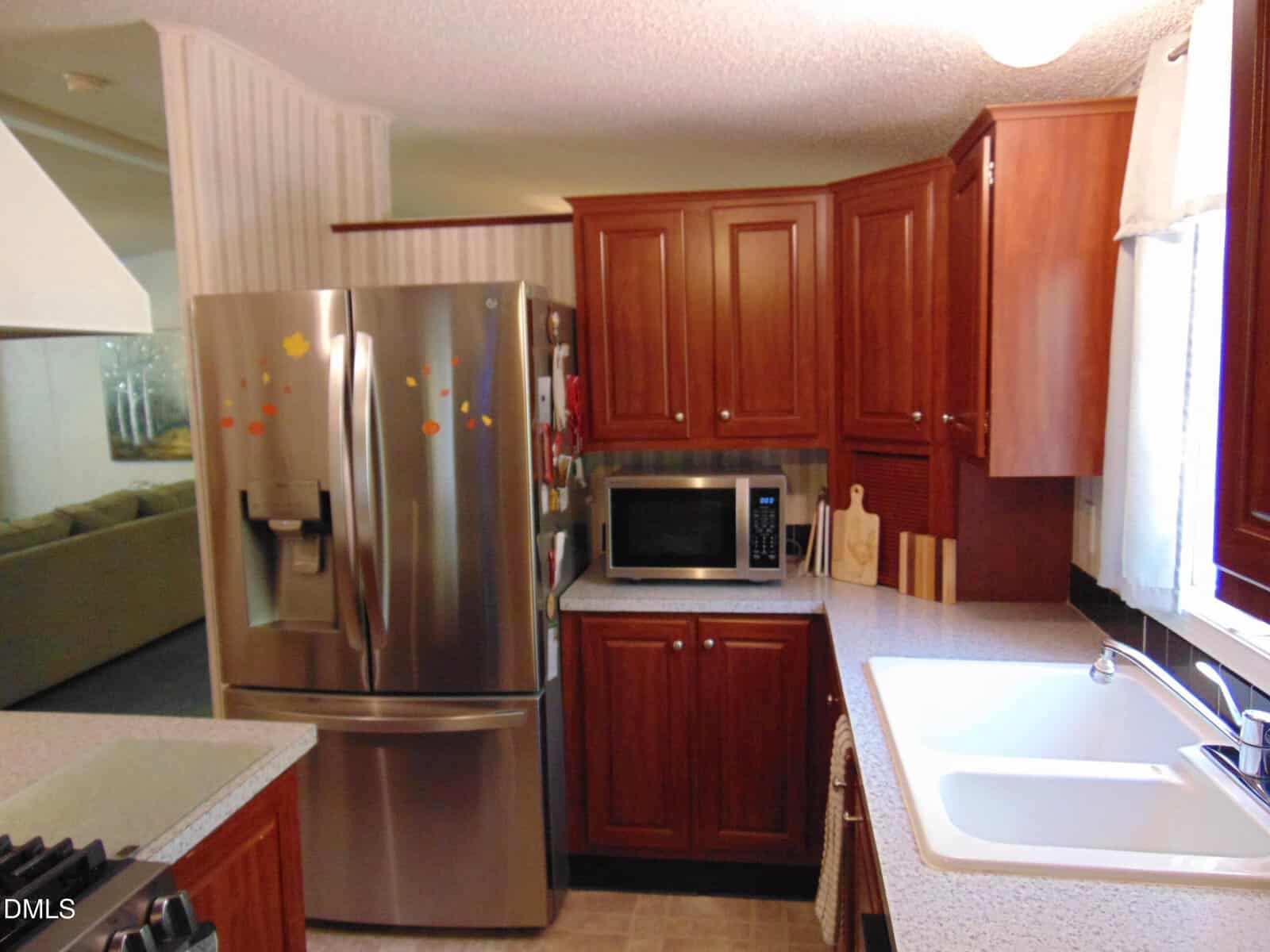 Small kitchen with stainless steel refrigerator, microwave, wooden cabinets, white countertop, sink under a window, and cutting boards on the counter. Living room visible in the background.