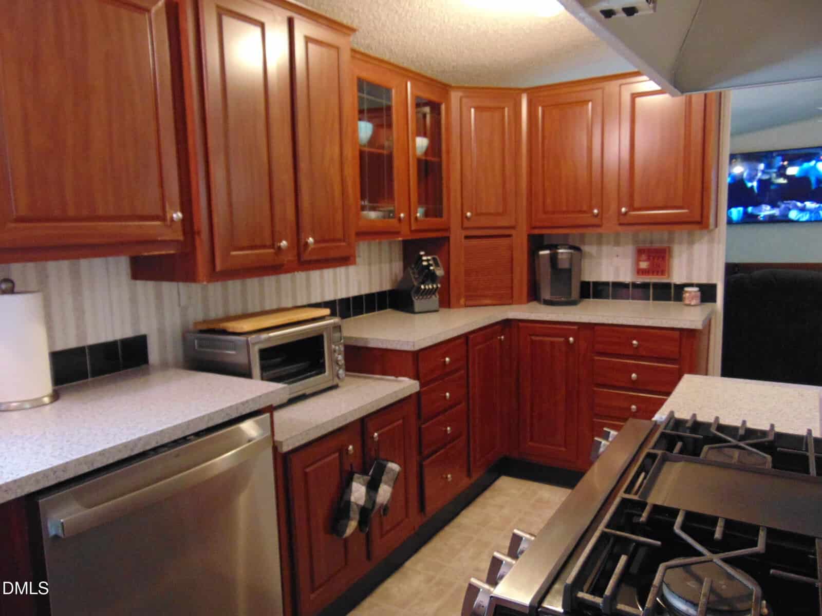A kitchen with wooden cabinets, stainless steel appliances, a gas stove, a toaster oven, a coffee maker, and light-colored countertops. A towel hangs on a drawer, and dishes are visible behind glass cabinet doors.