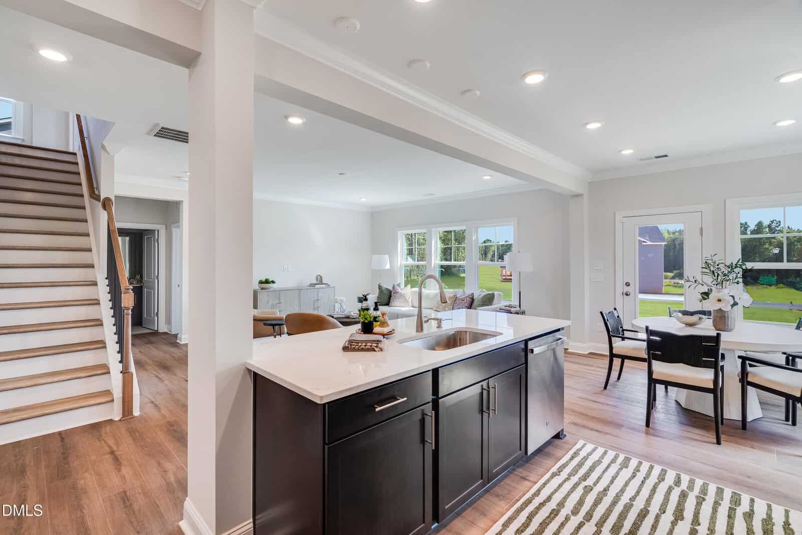 Modern open-concept kitchen with dark cabinets, white island, and stainless sink; adjacent dining area with table and chairs; living area with sofa; wood flooring; staircase to the left; large windows show green lawn outside.