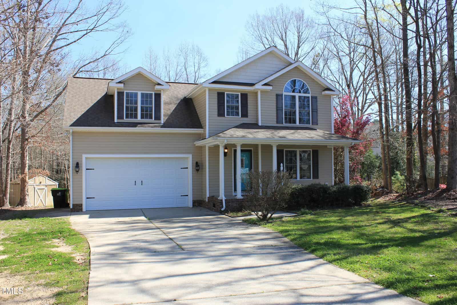 Two-story beige house with white trim, double garage, covered front porch, and arched window above the entrance. Leafless trees and a small storage shed are visible in the background. Concrete driveway in front.