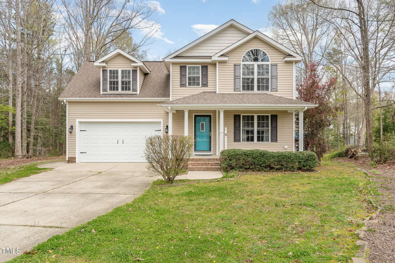 Two-story beige house with a teal front door, attached two-car garage, covered front porch, and a driveway. The yard has some grass, bushes, and a small tree. Tall trees surround the property in the background.