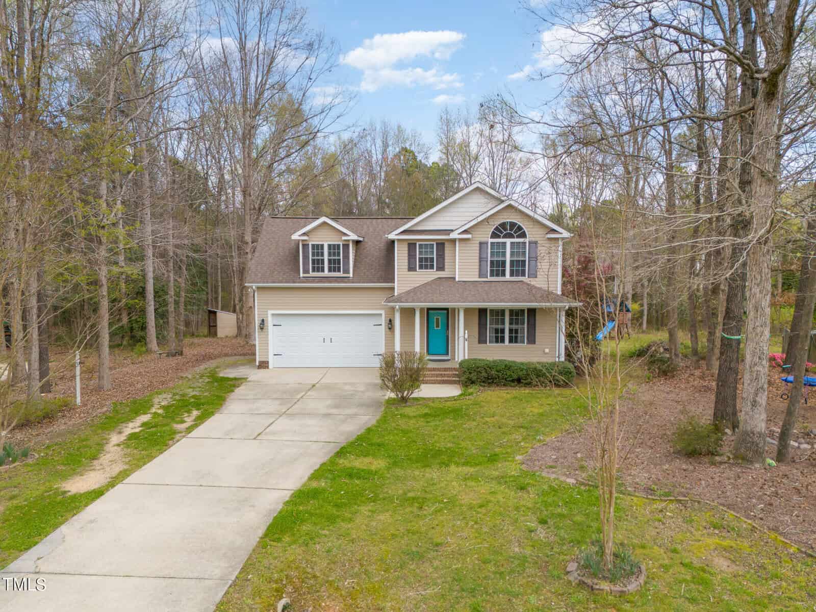 Two-story beige house at 60 Harley Court in Louisburg, featuring a teal front door, attached two-car garage, and concrete driveway, surrounded by trees and a lawn with sparse landscaping on a cloudy day.