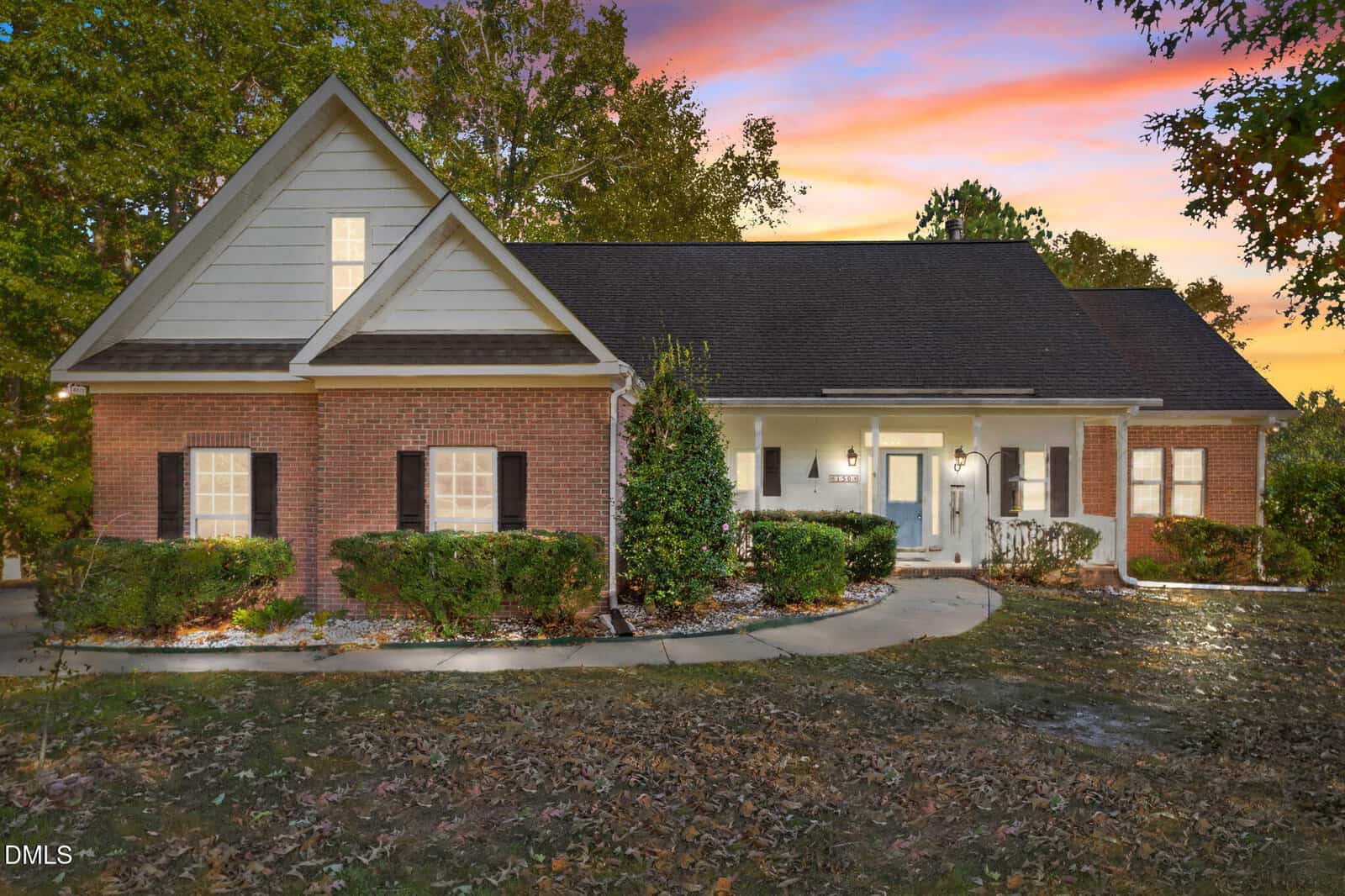 At sunset, 150 Williamston Ridge Drive in Youngsville features a brick and siding house with white trim and dark shutters, surrounded by trees and landscaping. The front entry glows warmly under the colorful pink and orange sky.