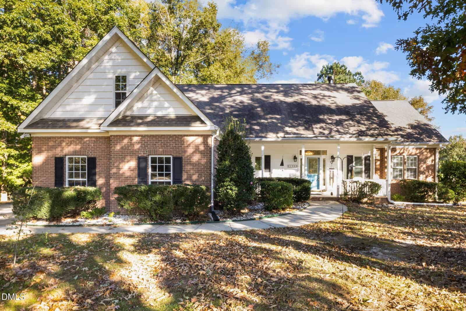 A single-story brick house with white siding accents, black shutters, and a front porch. The yard features bushes, trees, and fallen leaves, and a curved walkway leads to the front door.
