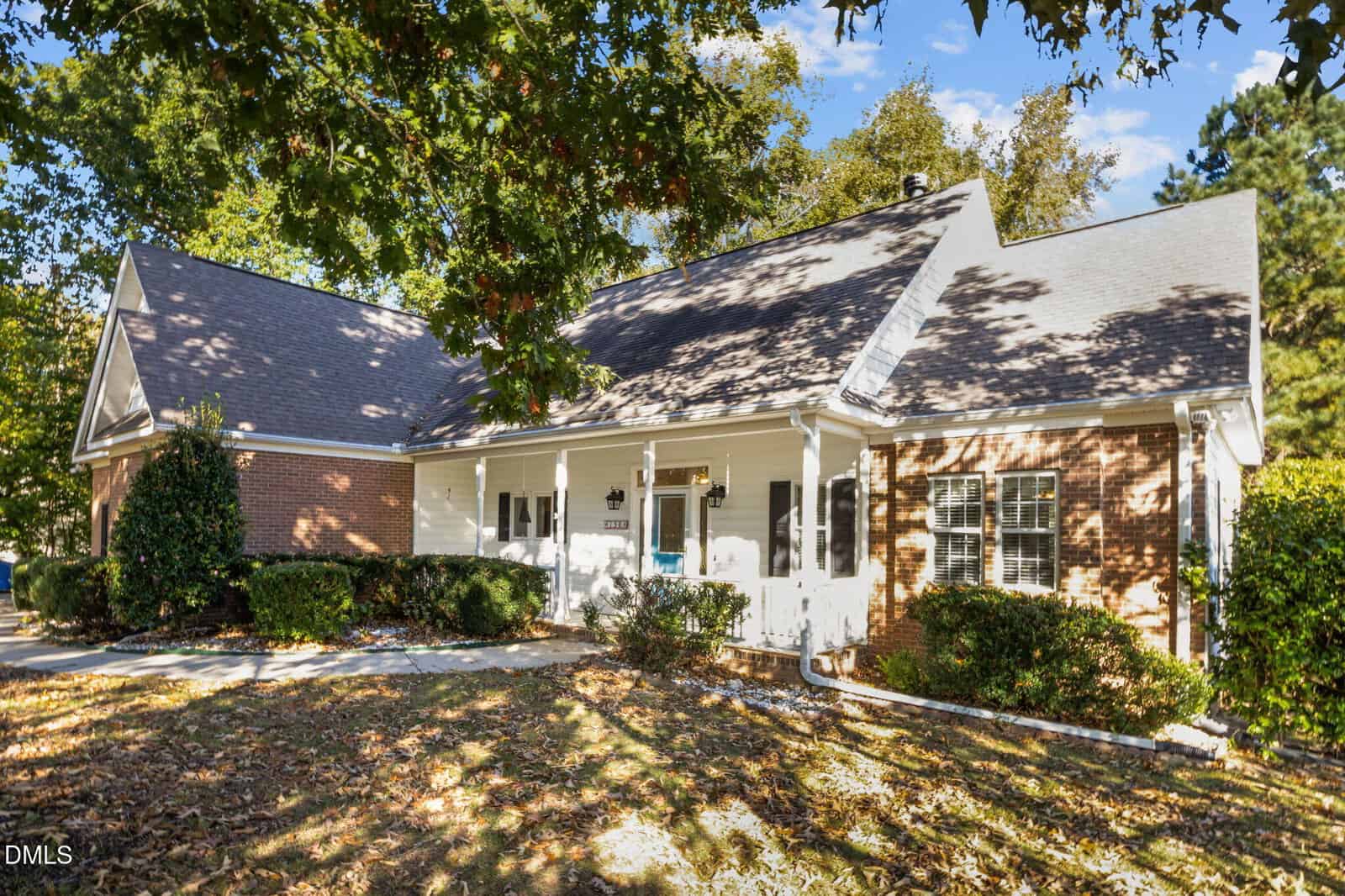 Single-story house with a brick and white siding exterior, front porch with white railings, surrounded by bushes and trees, with autumn leaves scattered on the lawn.