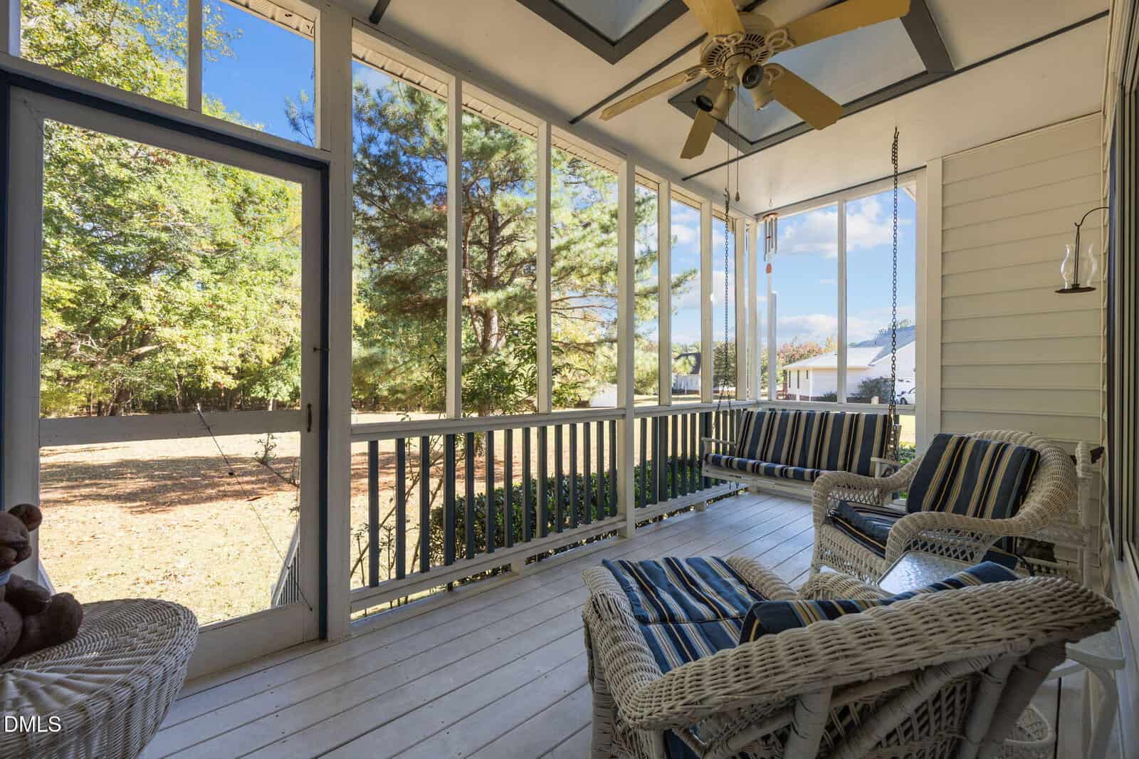 A screened-in porch with white wooden floors, wicker furniture with striped cushions, a ceiling fan, and views of a grassy yard and trees outside. Sunlight filters through the screens, creating a bright, airy atmosphere.