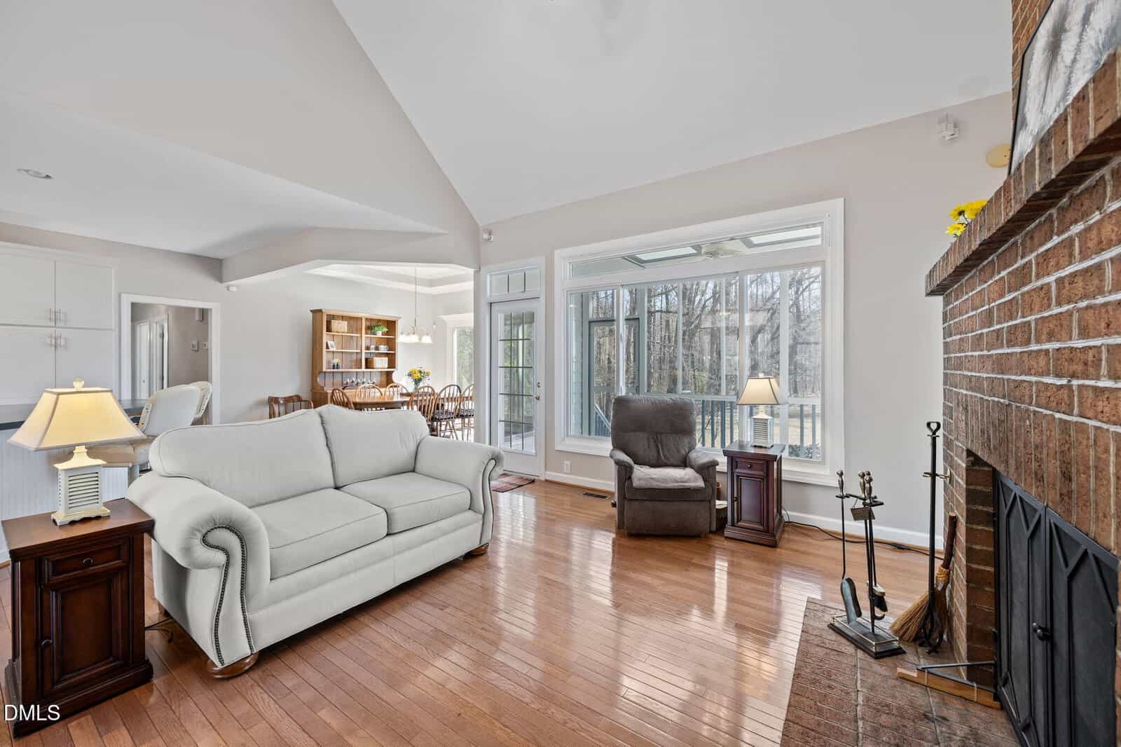 A living room with a white sofa, a brown armchair, and a brick fireplace. Hardwood floors, large windows, and a dining area with a wooden table and chairs are visible in the background.