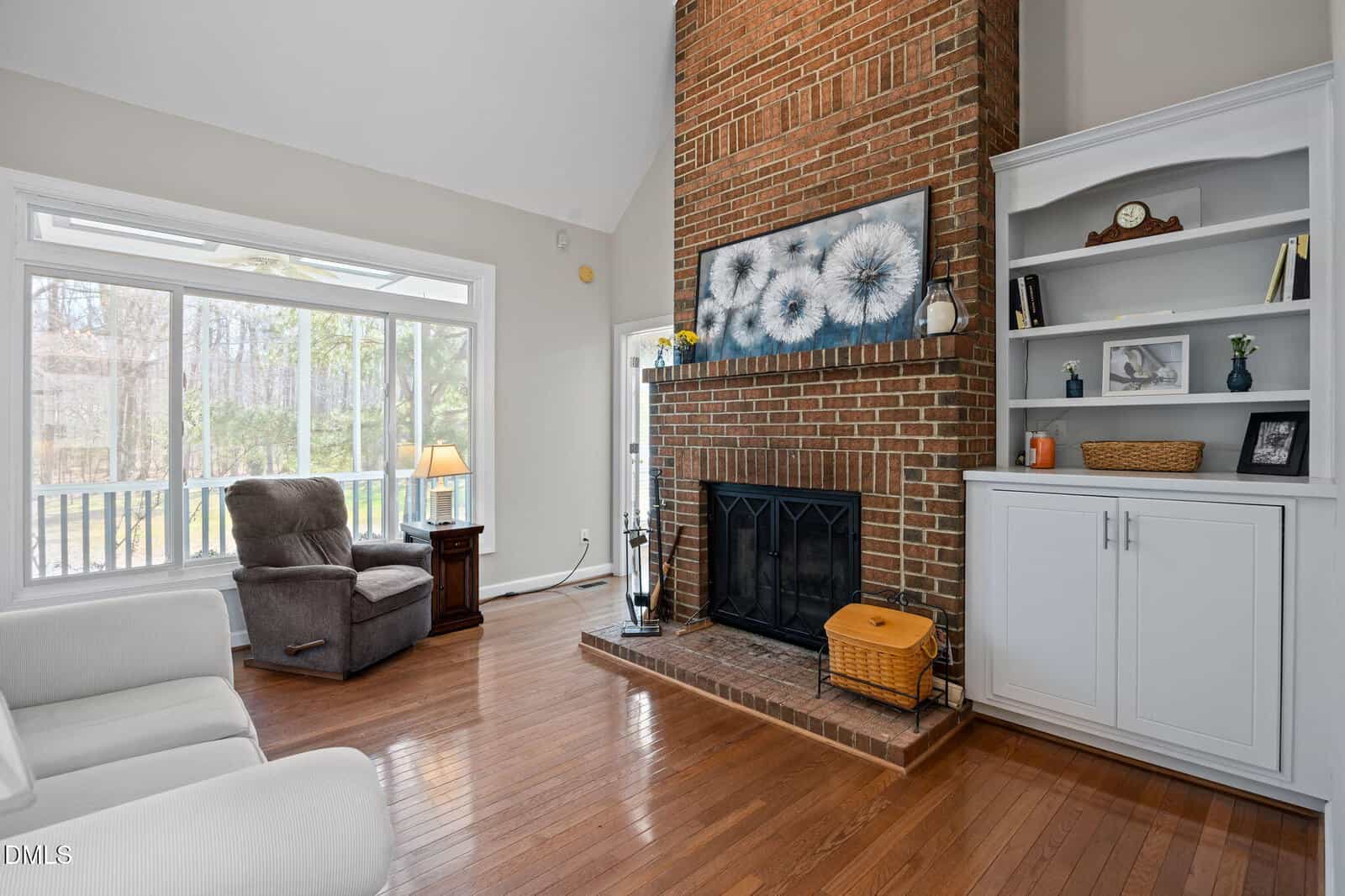 Bright living room with hardwood floors, large window, a brick fireplace, white built-in shelves, two armchairs, a side table with a lamp, and decorative wall art above the mantel.