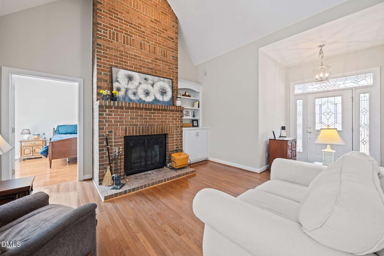 A living room with a tall brick fireplace, firewood, and fireplace tools. Above the mantel is a large floral painting. There are built-in shelves and cabinets, hardwood floors, and an entryway visible in the background.